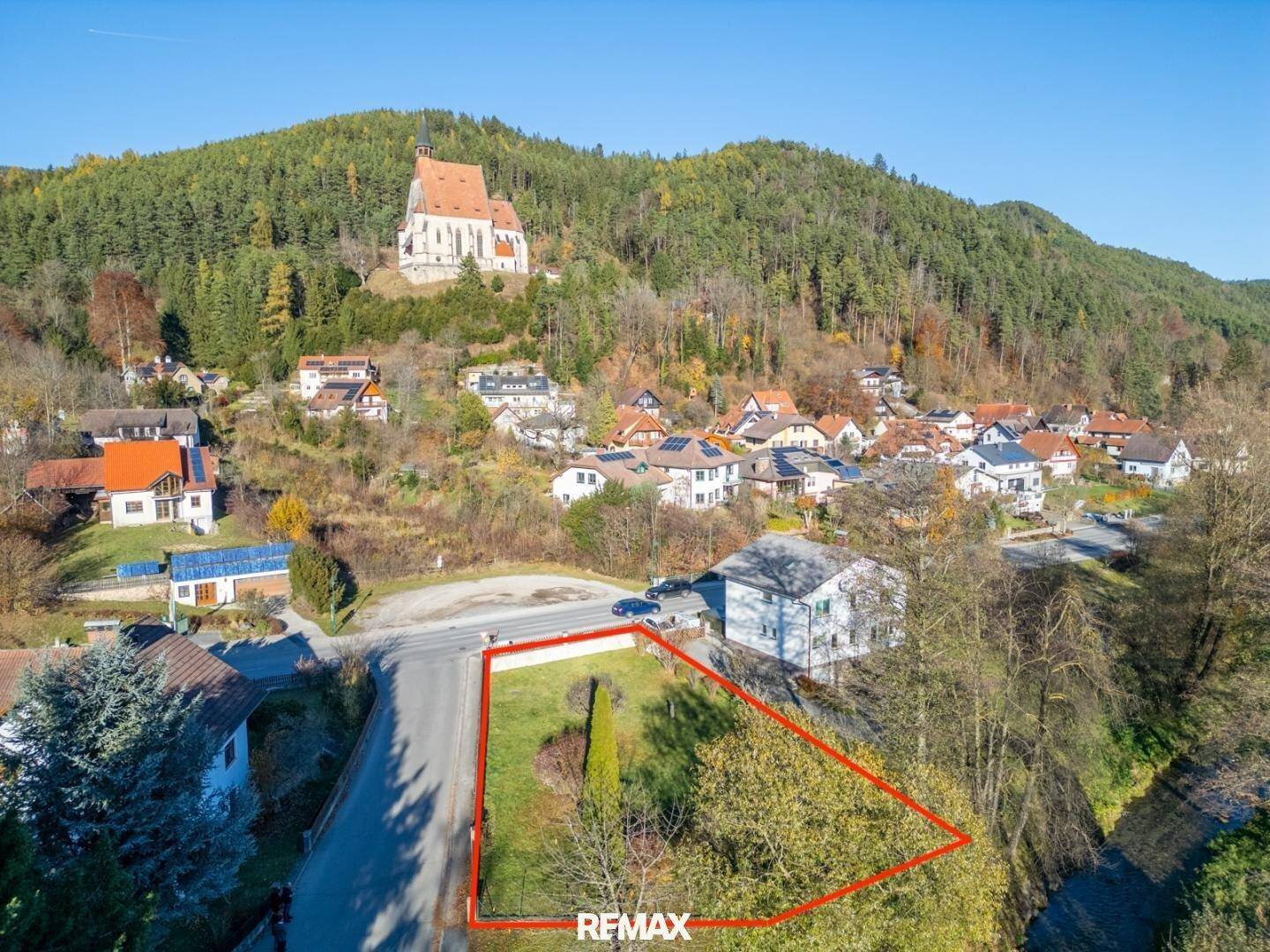 Luftaufnahme eines Grundstücks mit roter Umrandung, umgeben von Häusern und bewaldeten Hügeln mit einer Kirche.