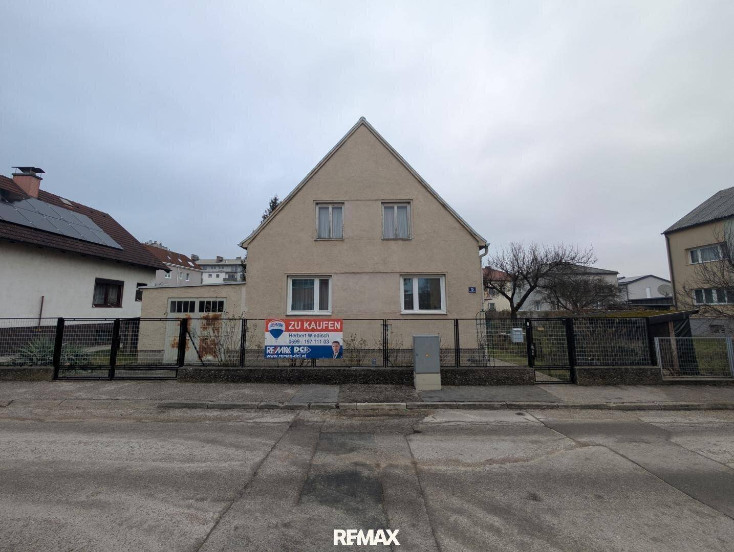 Vorderansicht des Einfamilienhauses von der Straße aus mit einem 'Zu Kaufen'-Schild an einem bewölkten Tag.