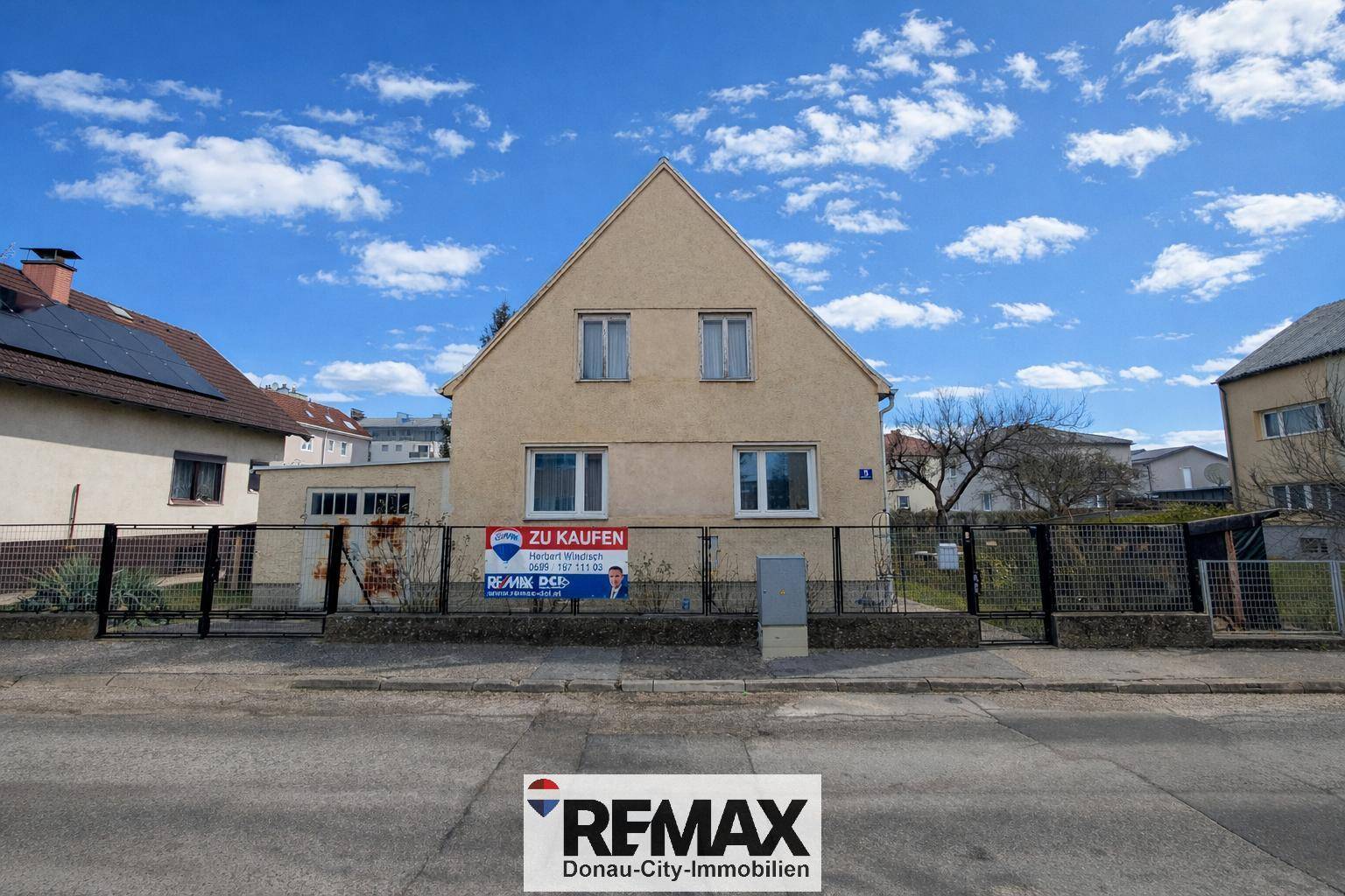 Vorderansicht eines Einfamilienhauses mit Zaun und Schild unter blauem Himmel.