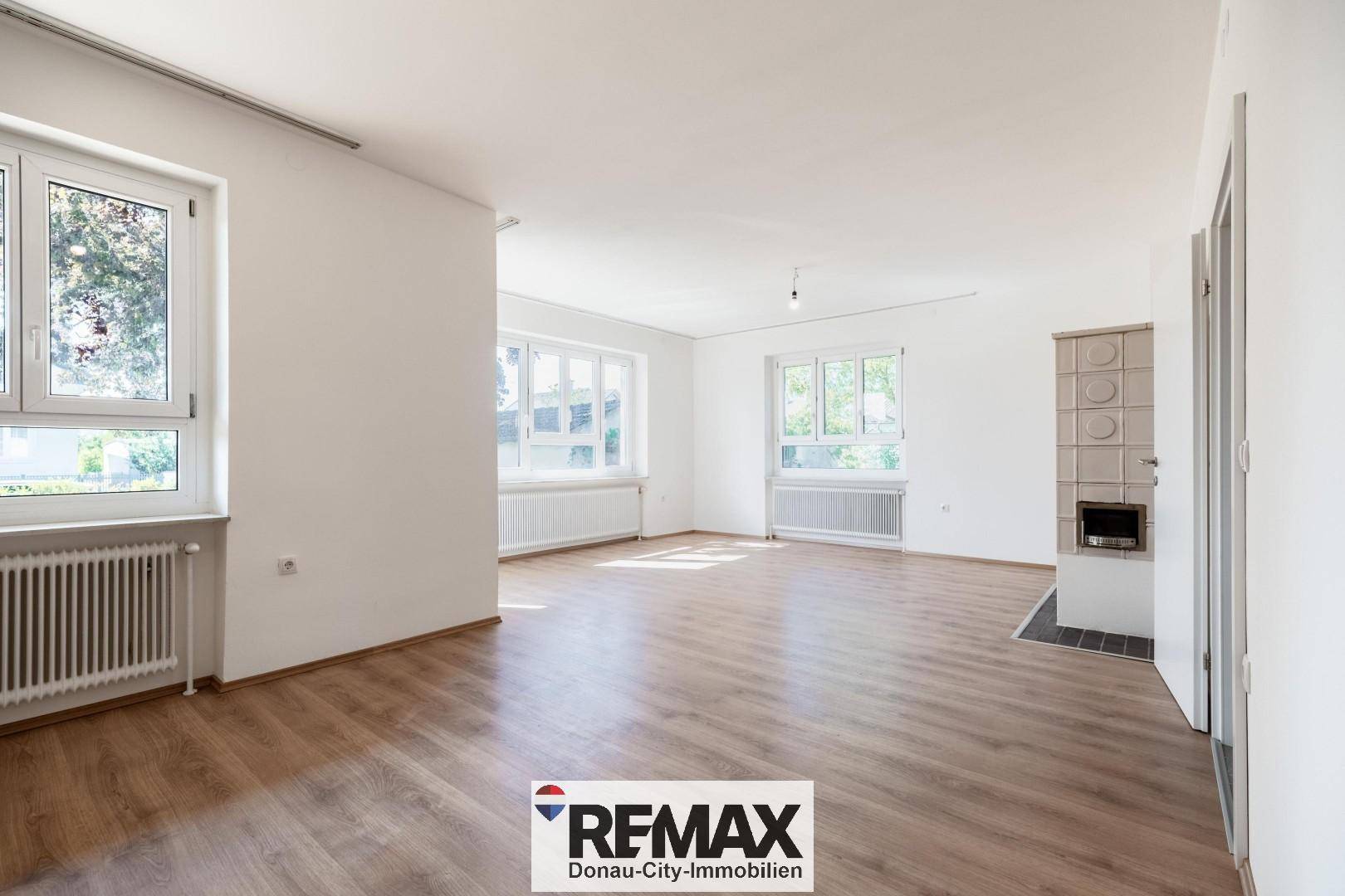 Geräumiges Wohnzimmer mit Laminatboden, großen Fenstern und einem Kachelofen.