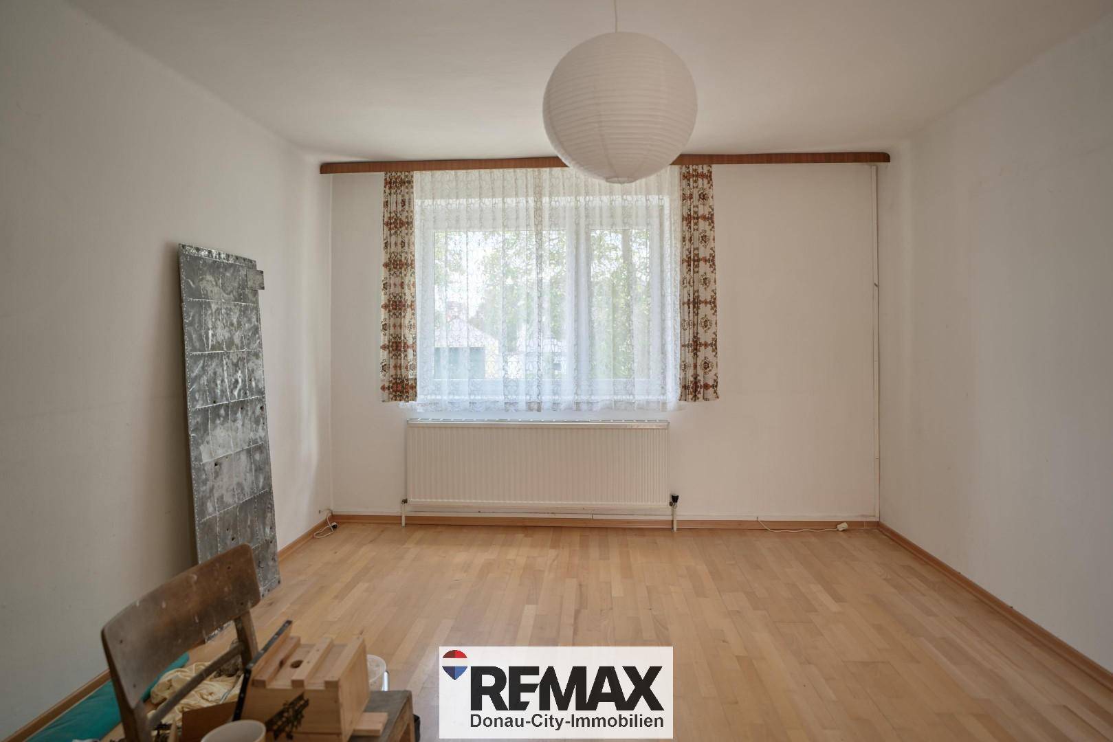 Leeres Zimmer mit Holzboden, großem Fenster und Heizkörper.
