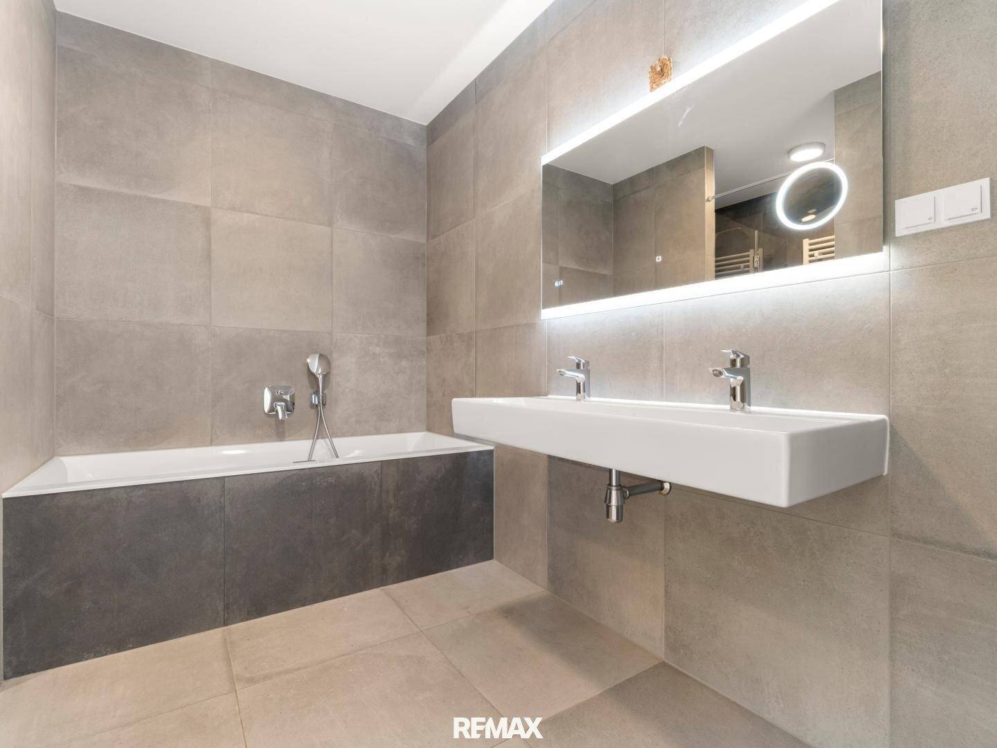 Modernes Badezimmer mit großen grauen Fliesen, Doppelwaschbecken und integrierter Badewanne.