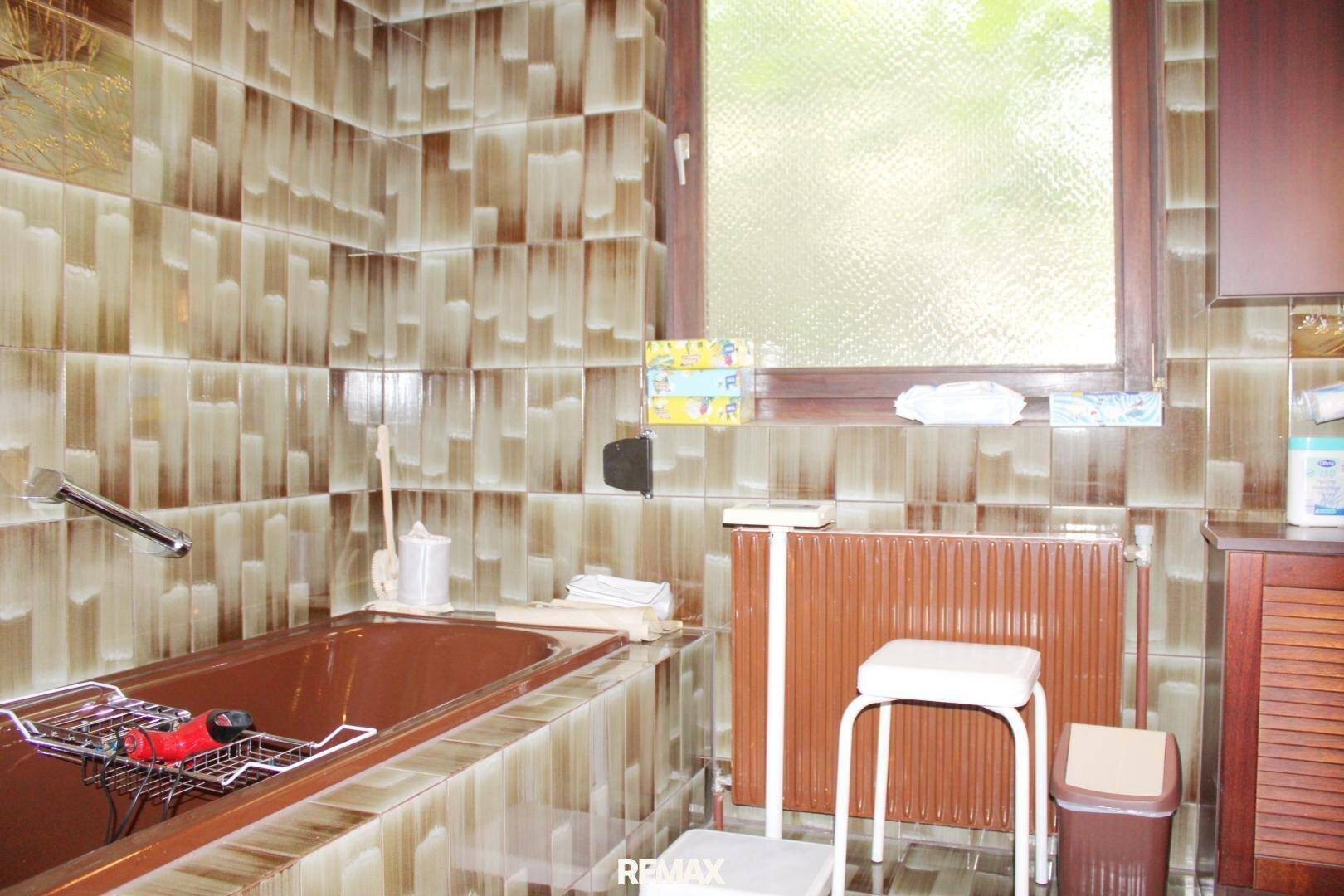 Badezimmer im 80er-Jahre-Stil mit brauner Badewanne, gemusterten Fliesen und Fenster.