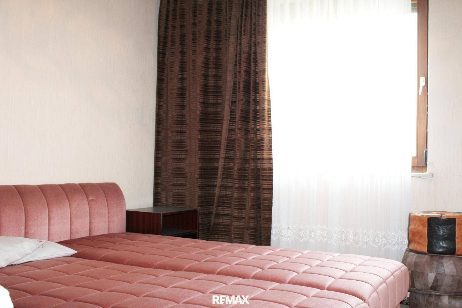Schlafzimmer mit rosa Bett, dunklen Vorhängen und einem Fenster, das viel Tageslicht hereinlässt.