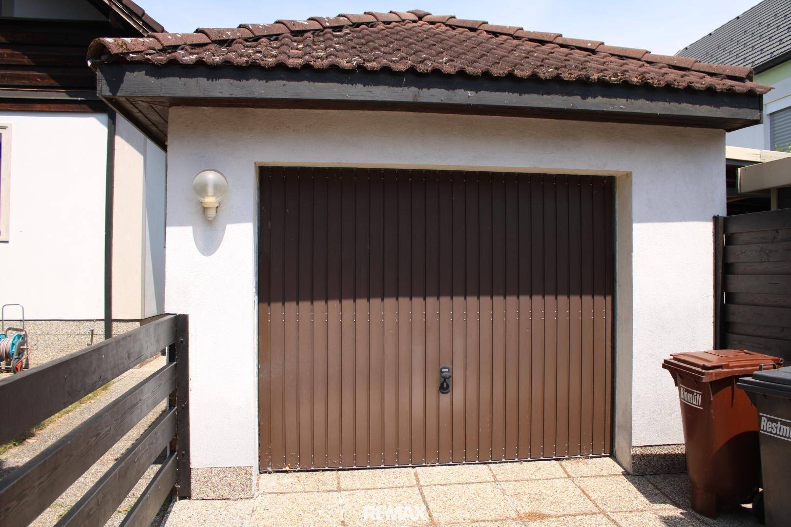Einzelgarage mit braunem Tor und gepflasterter Zufahrt, neben Mülltonnen.