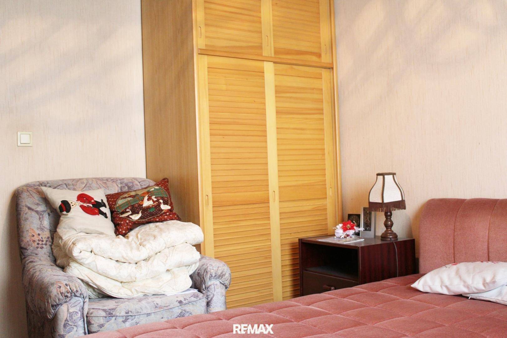 Schlafzimmer mit rosa Bett, Sessel und einem großen Holzschrank mit Lamellentüren.