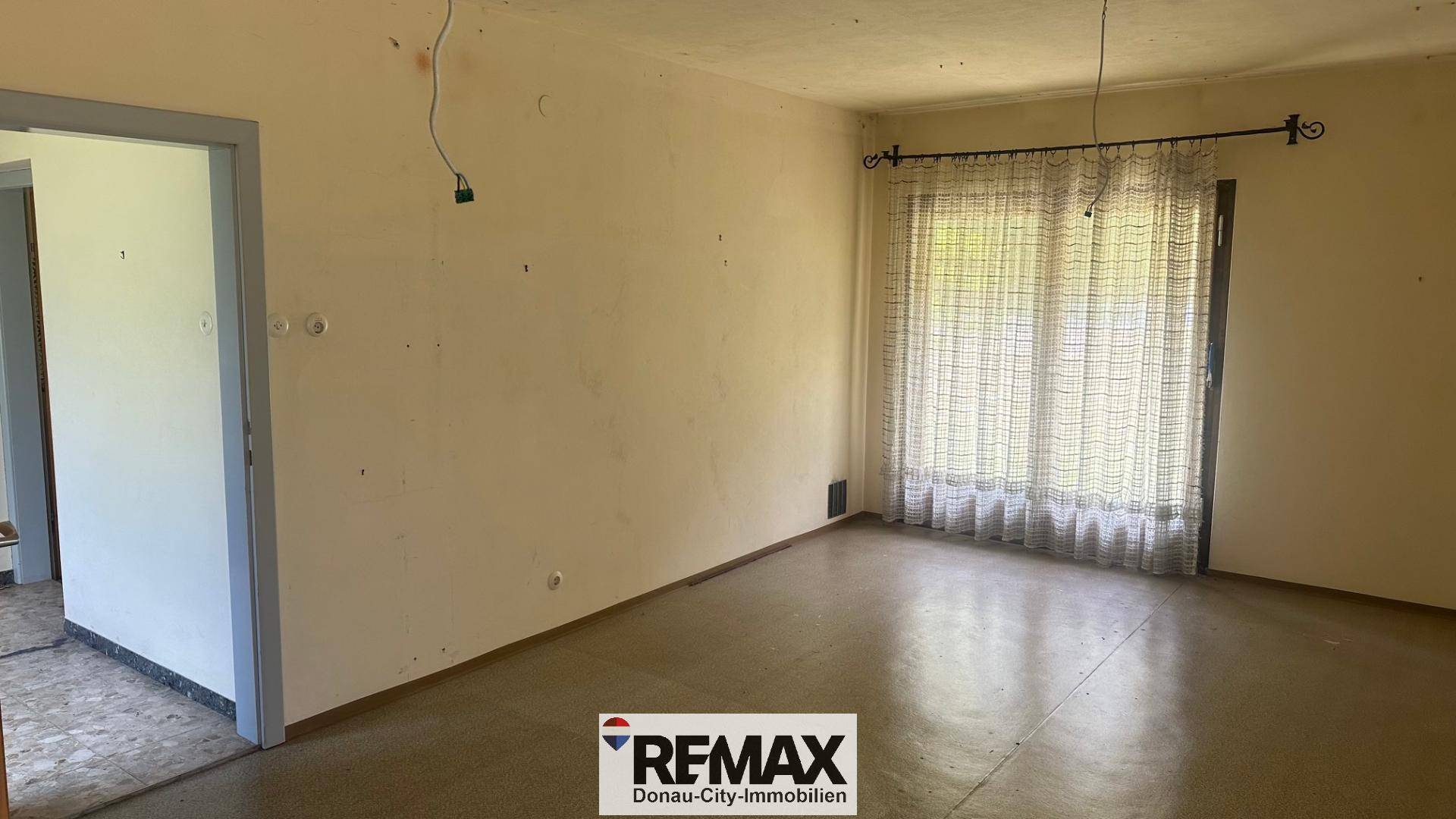 Leeres Wohnzimmer mit hellem Boden, großen Fenstern und Vorhängen, renovierungsbedürftig.