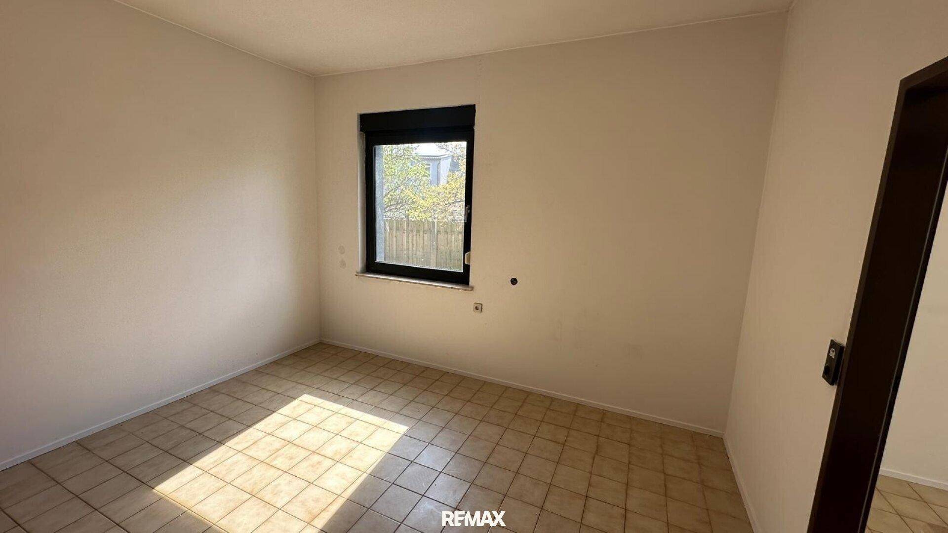 Helles Schlafzimmer mit Fliesenboden und einem Fenster mit Blick ins Grüne.