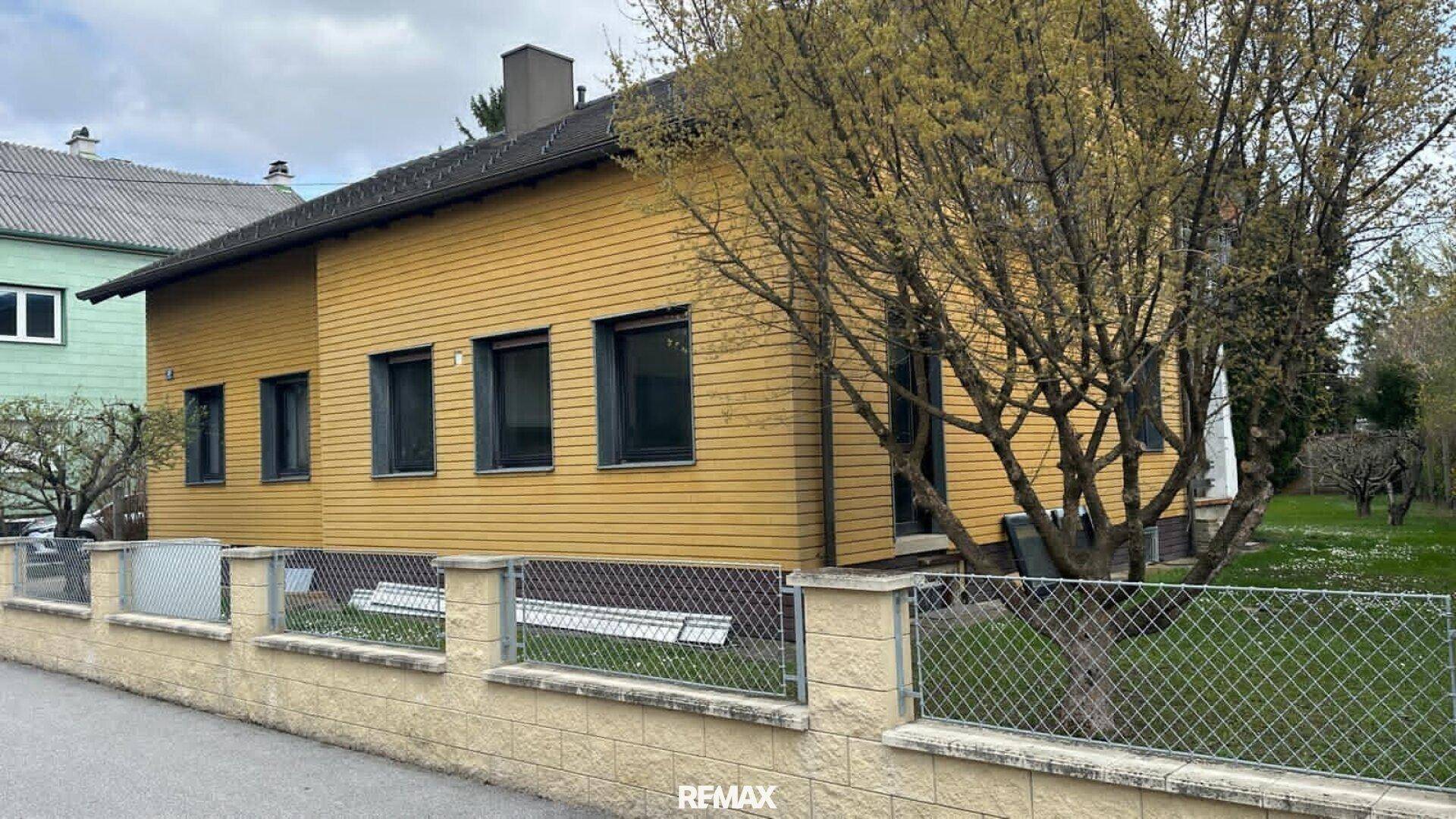 Vorderansicht des Hauses mit gelber Holzfassade und einem blühenden Baum im Vorgarten.