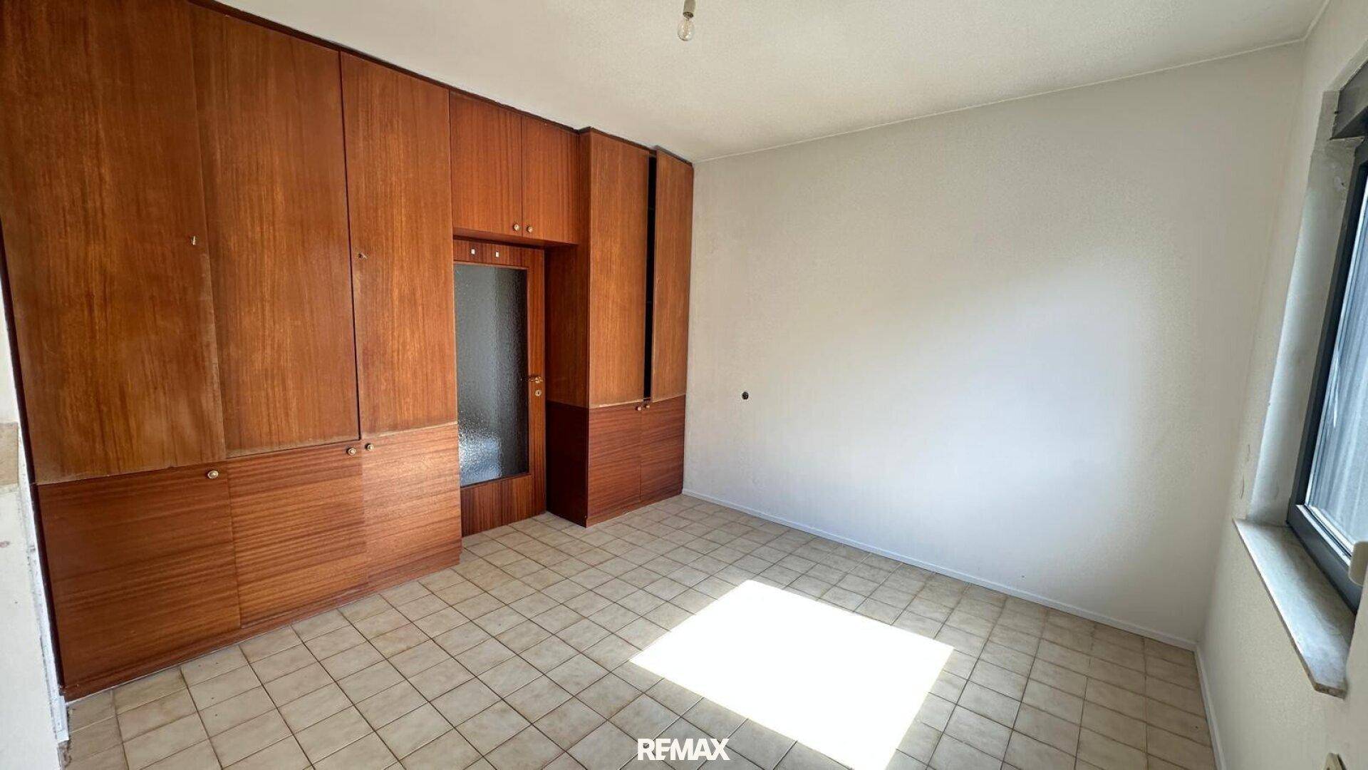 Schlafzimmer mit Einbauschränken, Fliesenboden und natürlichem Lichteinfall durch das Fenster.
