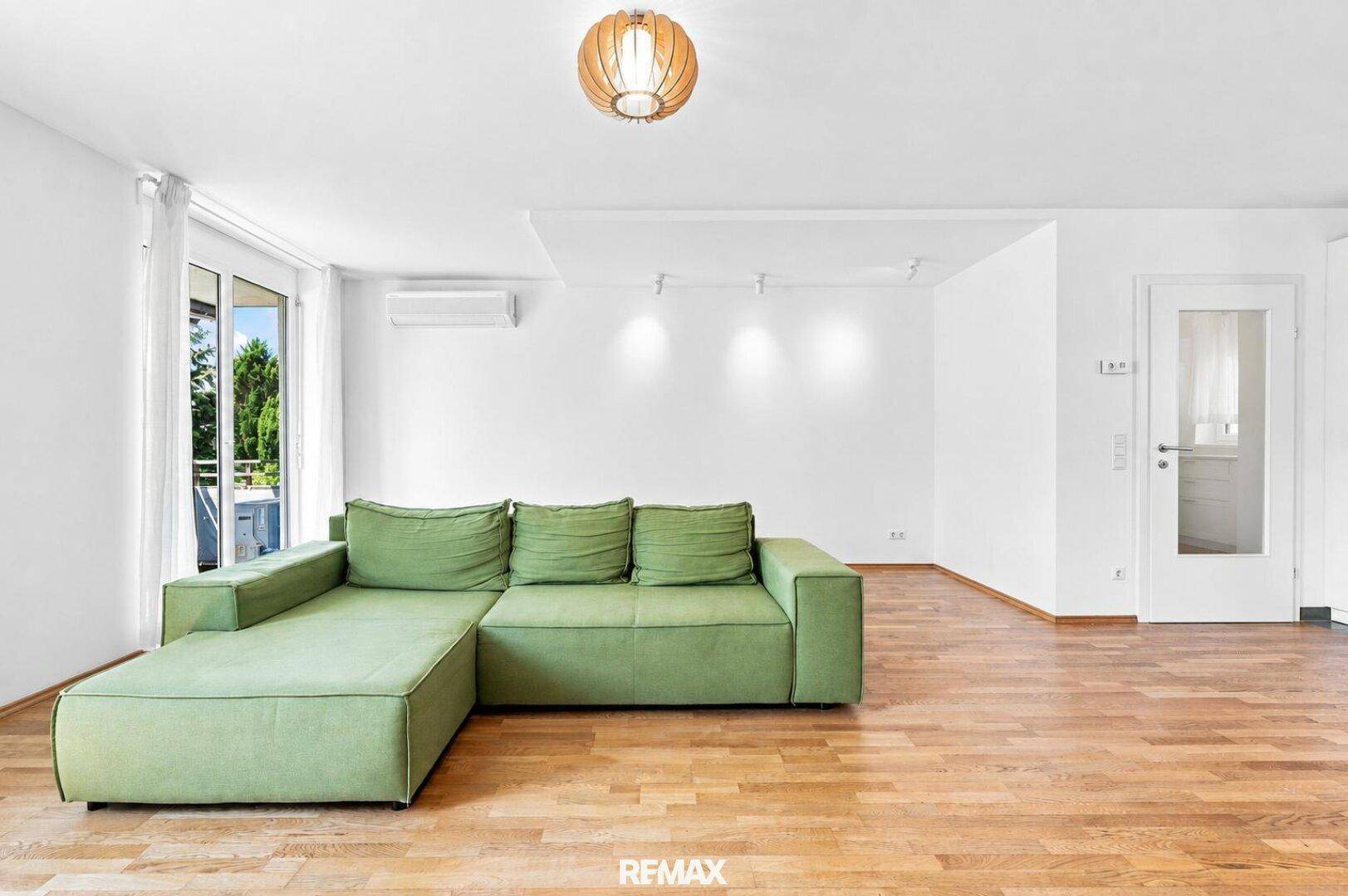 Lichtdurchflutetes Wohnzimmer mit Zugang zum Balkon, grüner Couch und Holzboden.