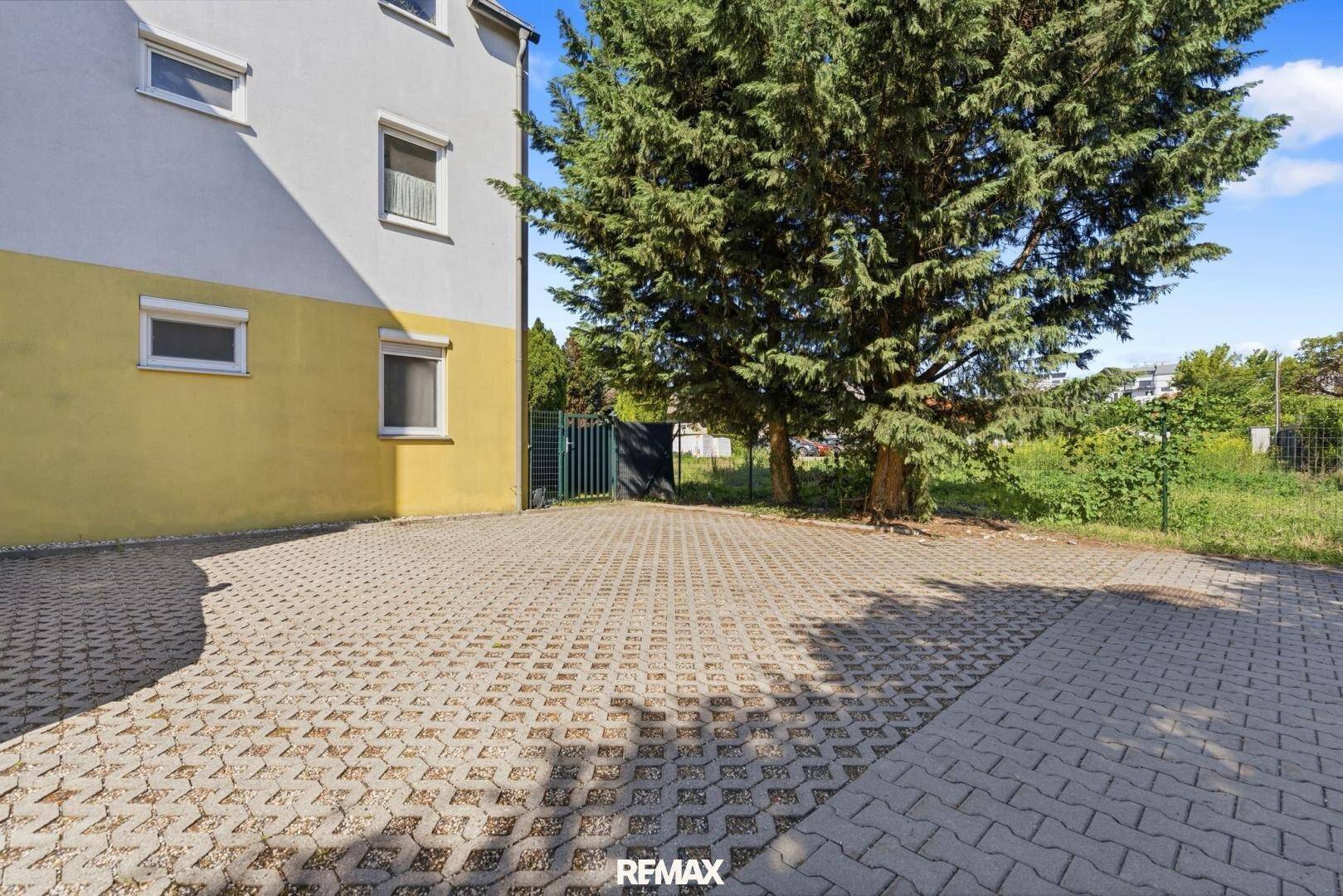 Außenbereich der Wohnanlage mit gepflastertem Parkplatz und altem Baumbestand unter blauem Himmel.