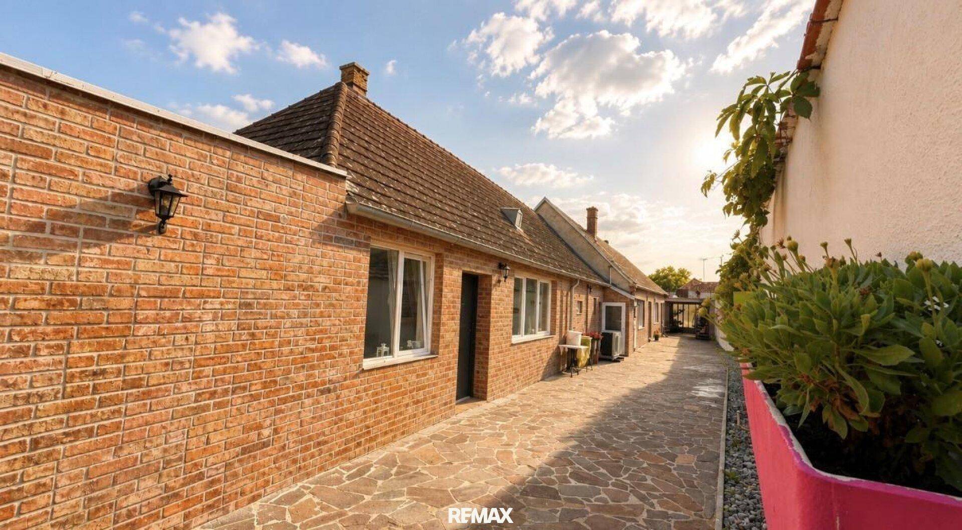 Lange gepflasterte Gasse entlang der Ziegelmauer des Hauses unter sonnigem Himmel.