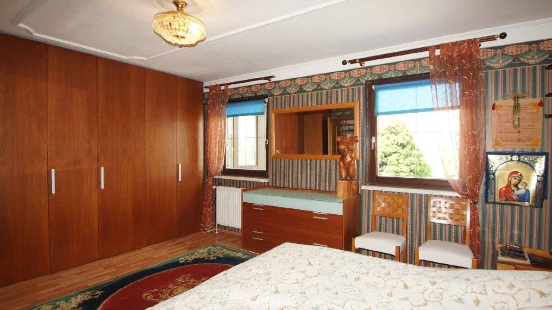 Schlafzimmer mit großem Holzschrank, gemusterter Tapete und zwei Fenstern mit Blick ins Grüne.