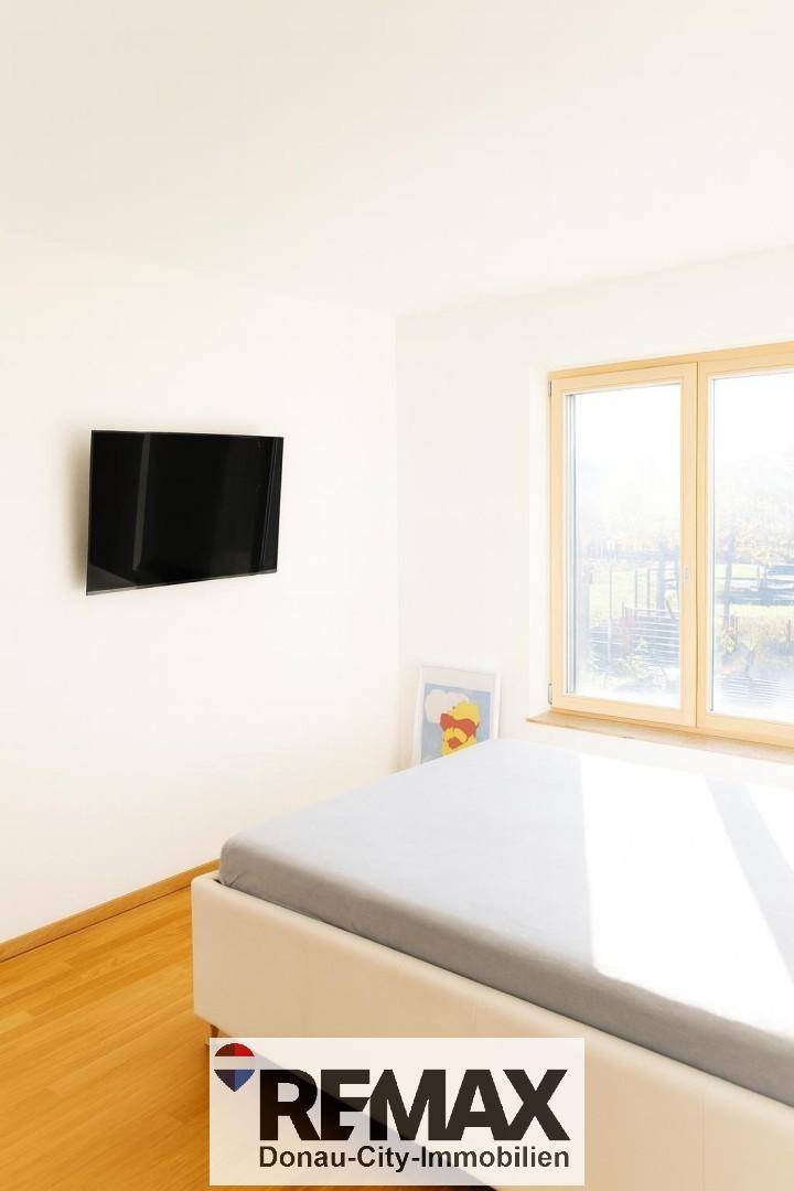 Schlafzimmer mit hellem Holzboden, großem Fenster und Wandfernseher.