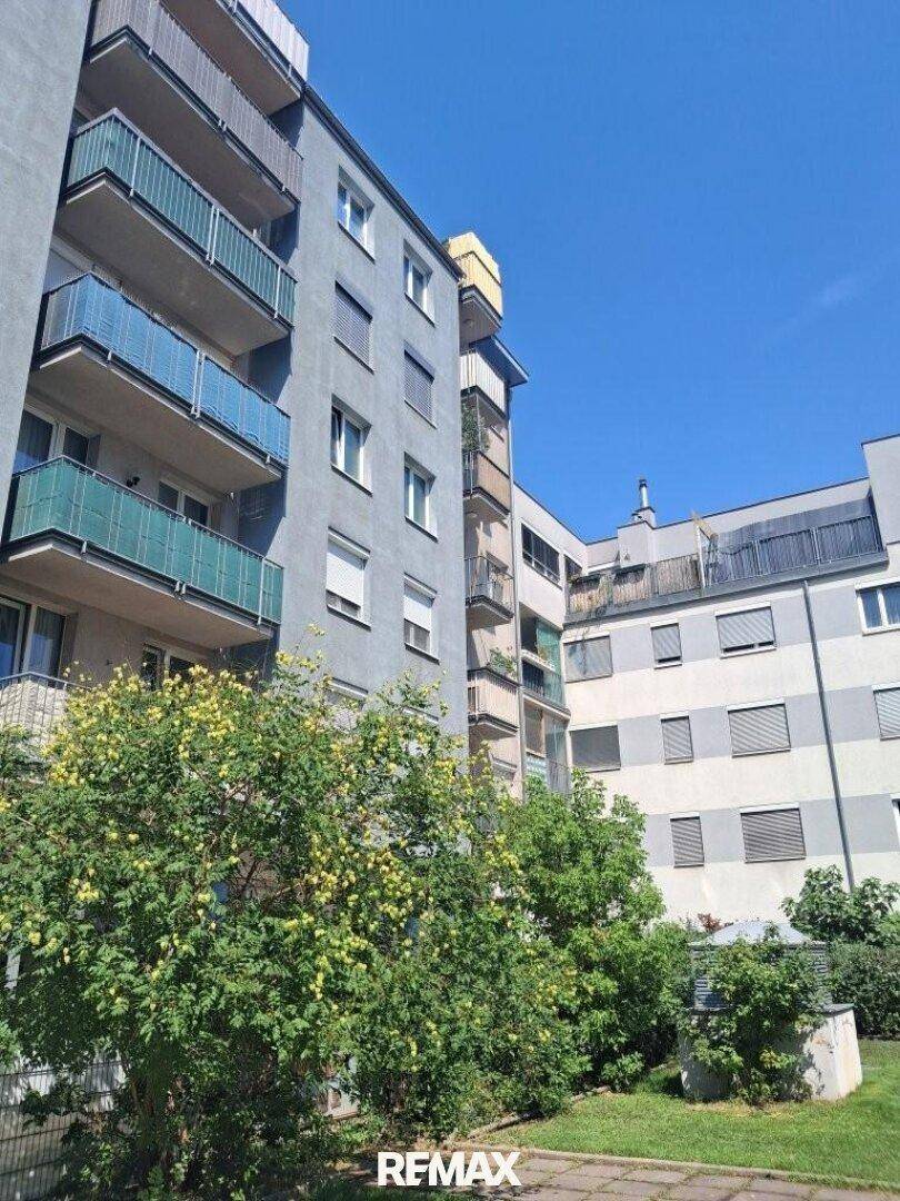 Mehrstöckiges Wohngebäude mit Balkonen und grünen Büschen im Vordergrund unter blauem Himmel.