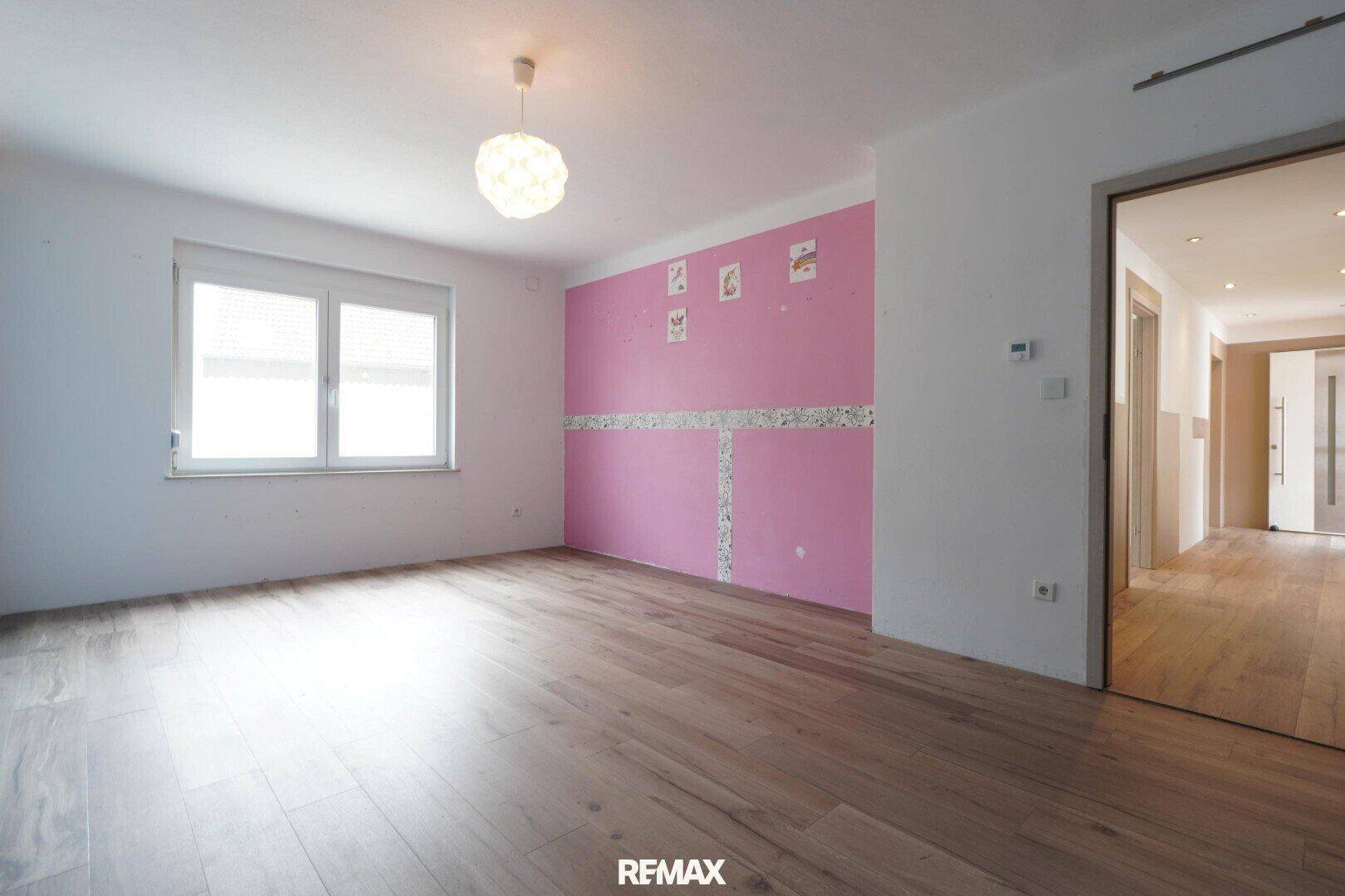 Kinderzimmer mit hellem Holzfußboden, einer rosa Akzentwand und einem großen Fenster.
