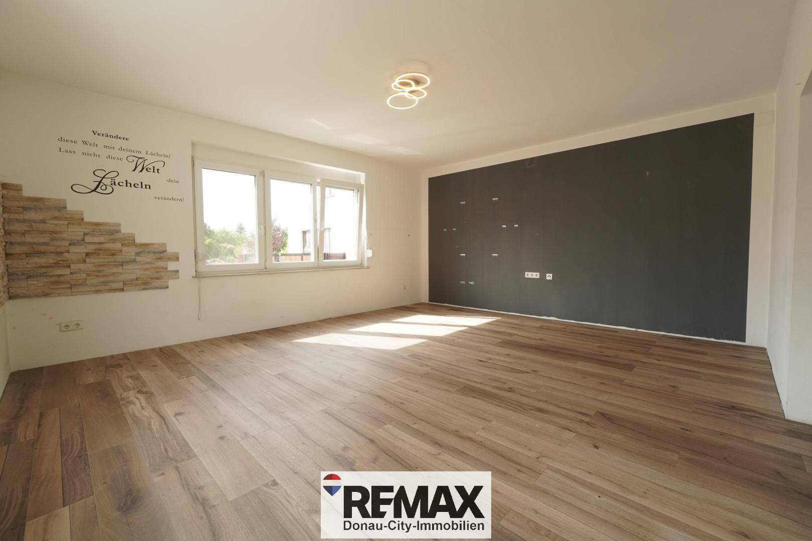 Helles Wohnzimmer mit großen Fenstern, Holzboden und einer dunklen Akzentwand.
