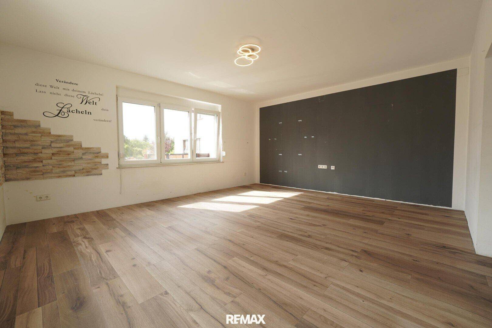Helles Wohnzimmer mit Holzfußboden, einer dunklen Akzentwand und viel Tageslicht durch große Fenster.