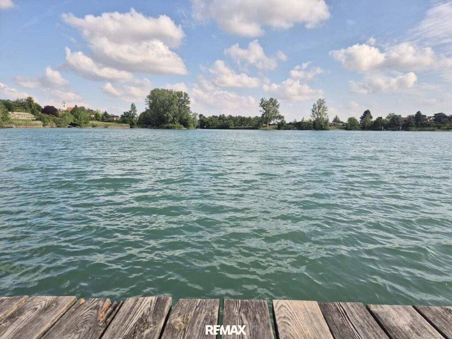Weitläufiger Blick über den See von einem Holzsteg unter einem bewölkten Himmel.