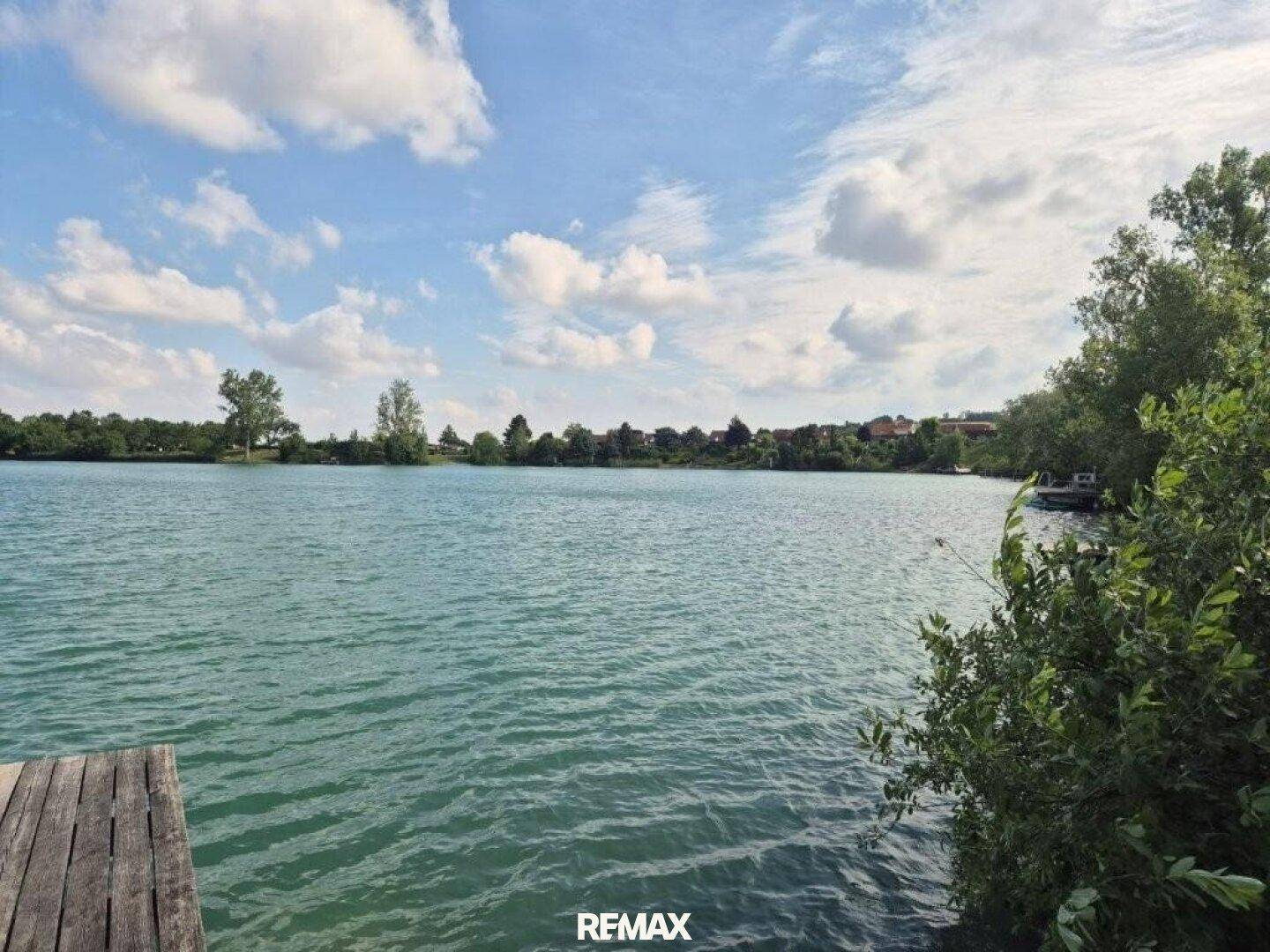 Panoramablick auf den See von einem Holzsteg mit Uferbewuchs am rechten Rand.