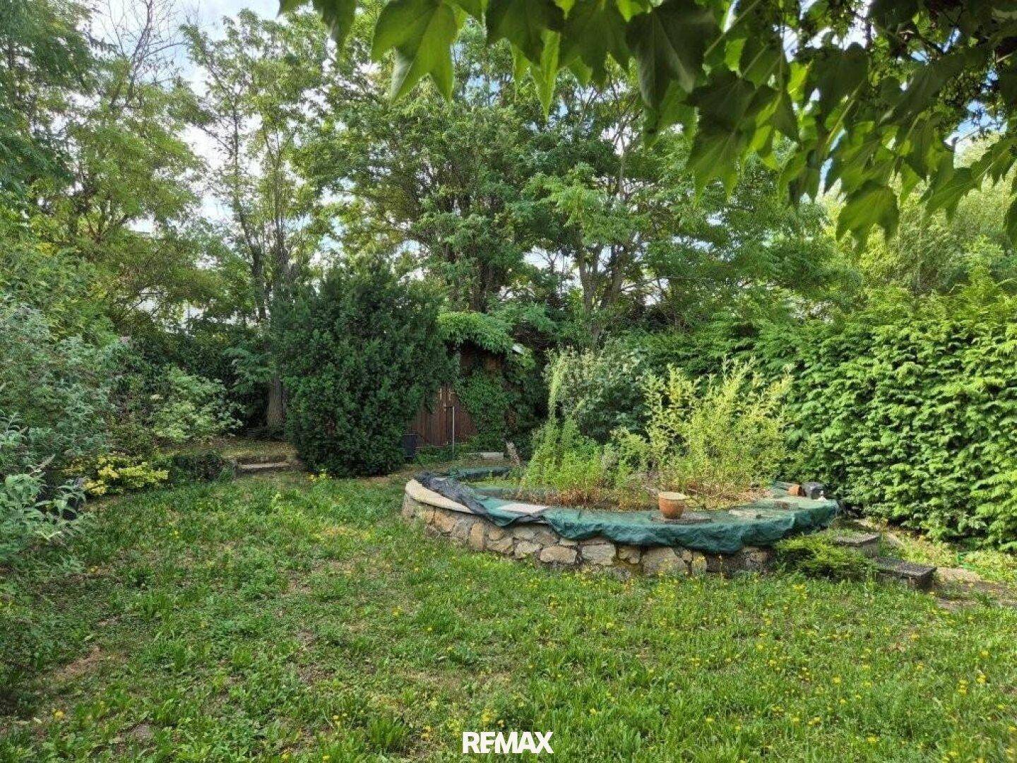 Naturbelassener Garten mit einem runden Teich oder Blumenbeet und altem Baumbestand.