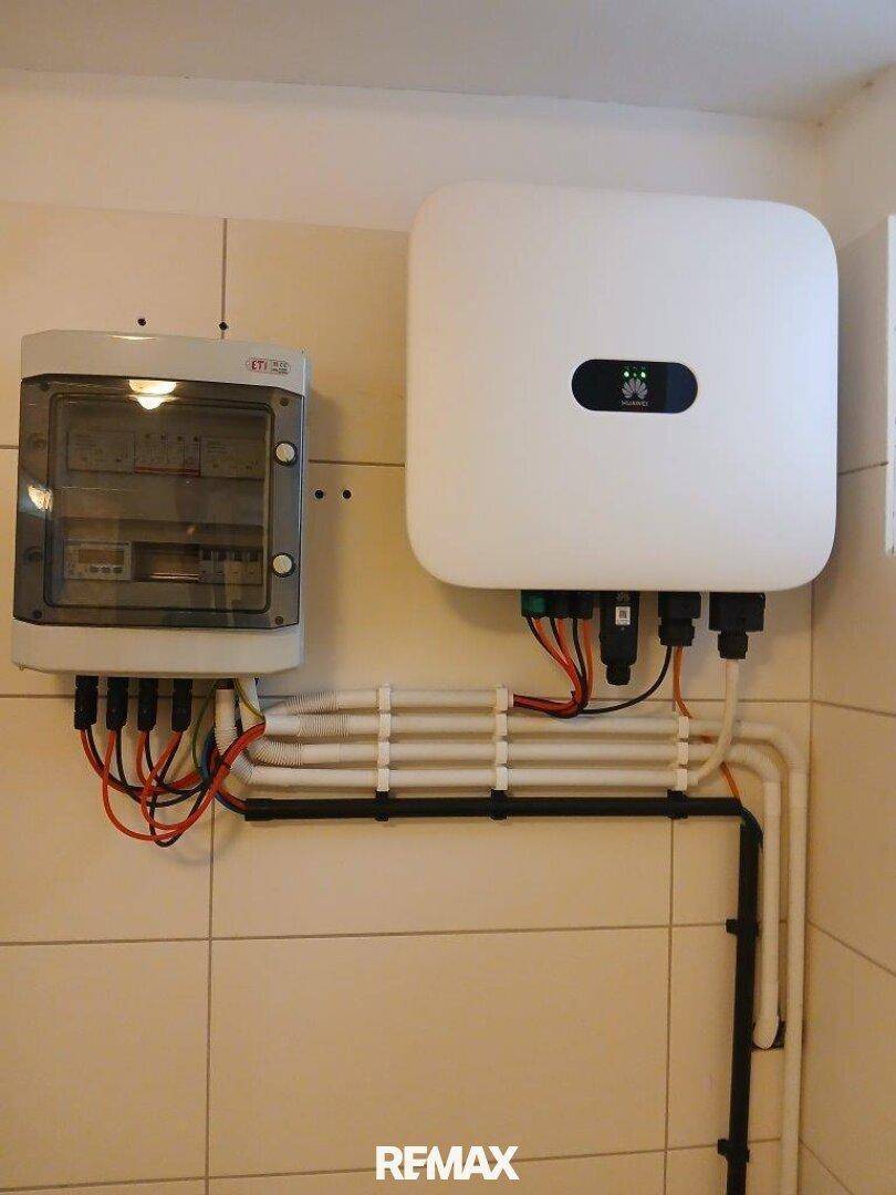Elektrische Installationen mit Sicherungskasten und Wechselrichter an einer gefliesten Wand im Technikraum.