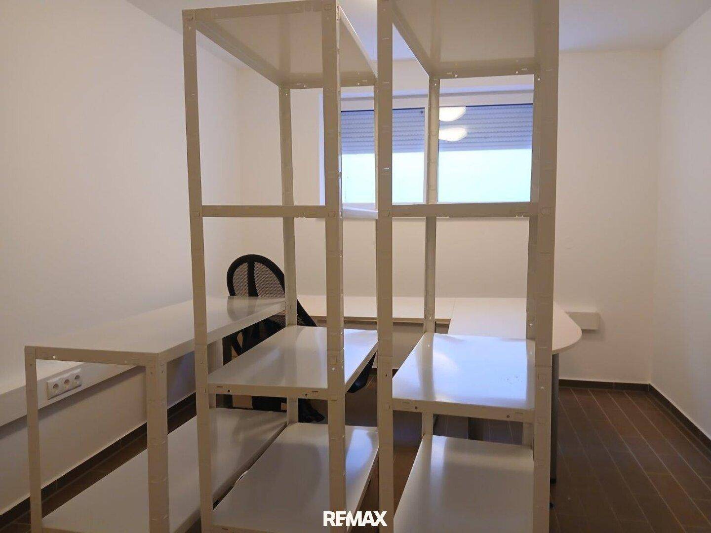 Helles Büro mit Regalsystem, Schreibtischen und einem Fenster mit Rollladen für natürliche Belichtung.
