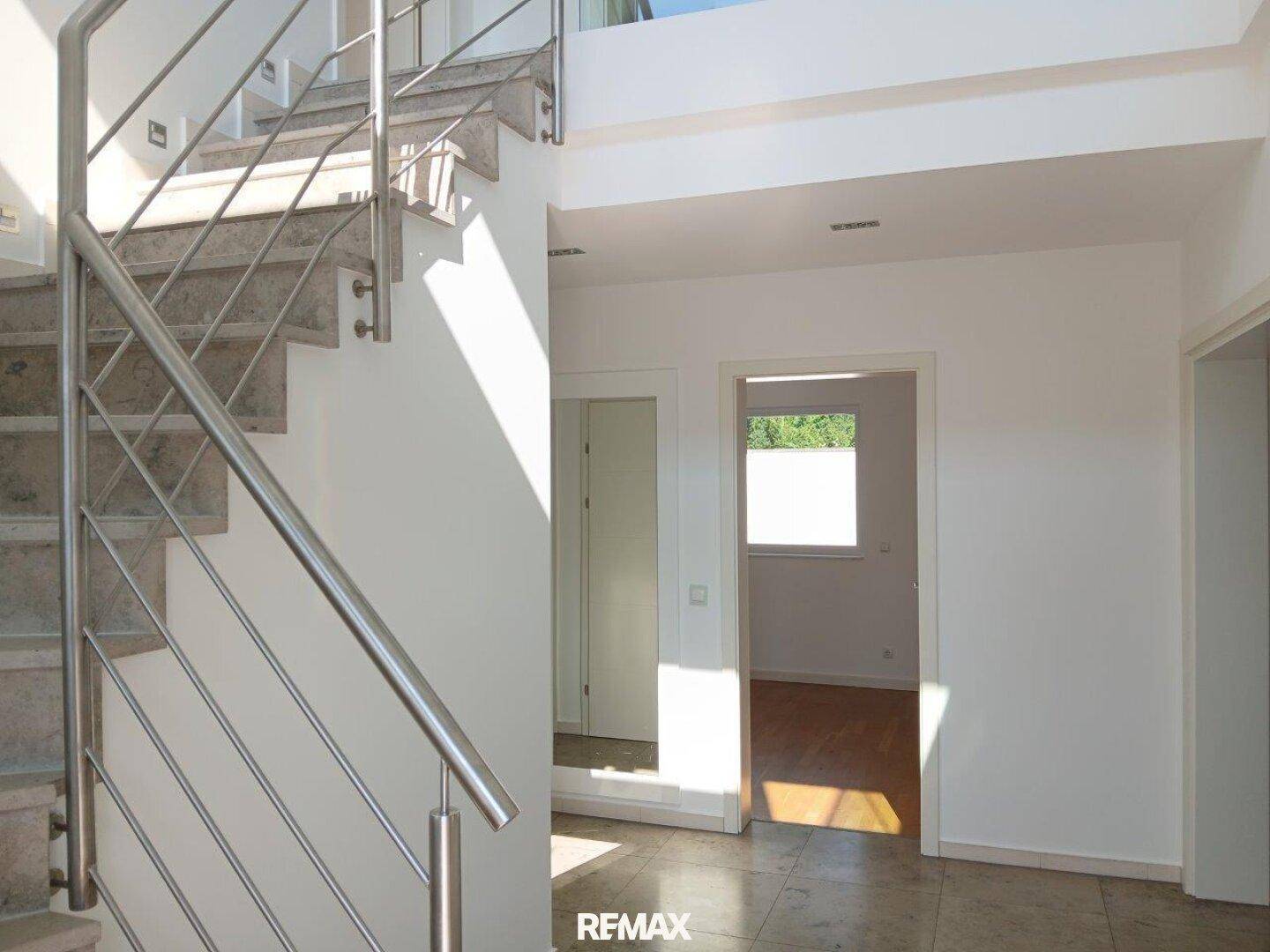 Heller Flur mit moderner Steintreppe und Edelstahlgeländer, durchflutet von Sonnenlicht.