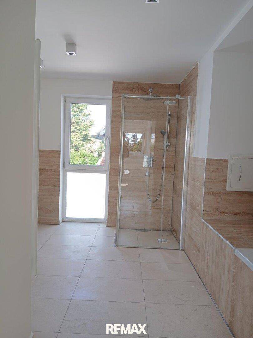 Modernes Badezimmer mit begehbarer Dusche, hellen Fliesen und Fenster.