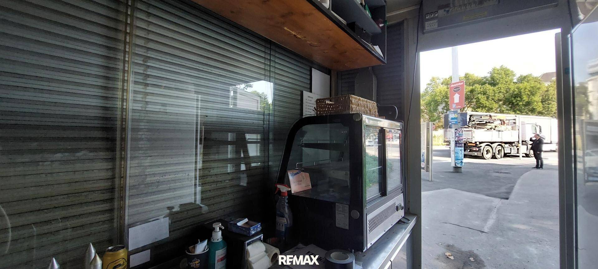 Blick in den Kiosk mit einer Kühlvitrine, Holzregal und geöffnetem Rollladen zur Straße.