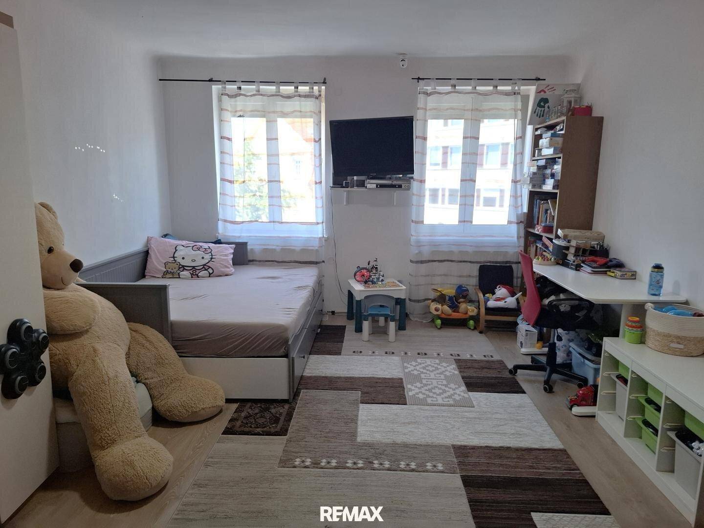 Kinderzimmer mit Bett, Schreibtisch, Spielzeug und zwei Fenstern für viel Tageslicht.