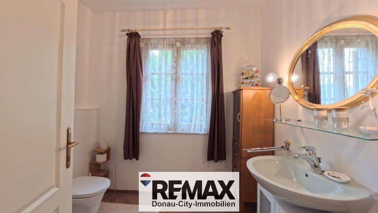 Helles Badezimmer mit Fenster, ovalem Spiegel und Einzelwaschbecken, das eine funktionale Einrichtung bietet.
