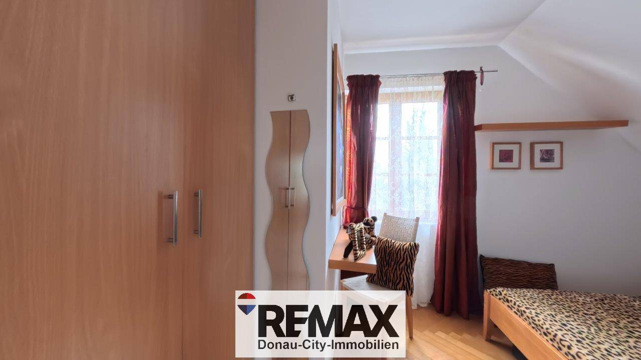 Schlafzimmer im Dachgeschoss mit Holzschrank und Fenster mit roten Vorhängen, die Gemütlichkeit ausstrahlen.