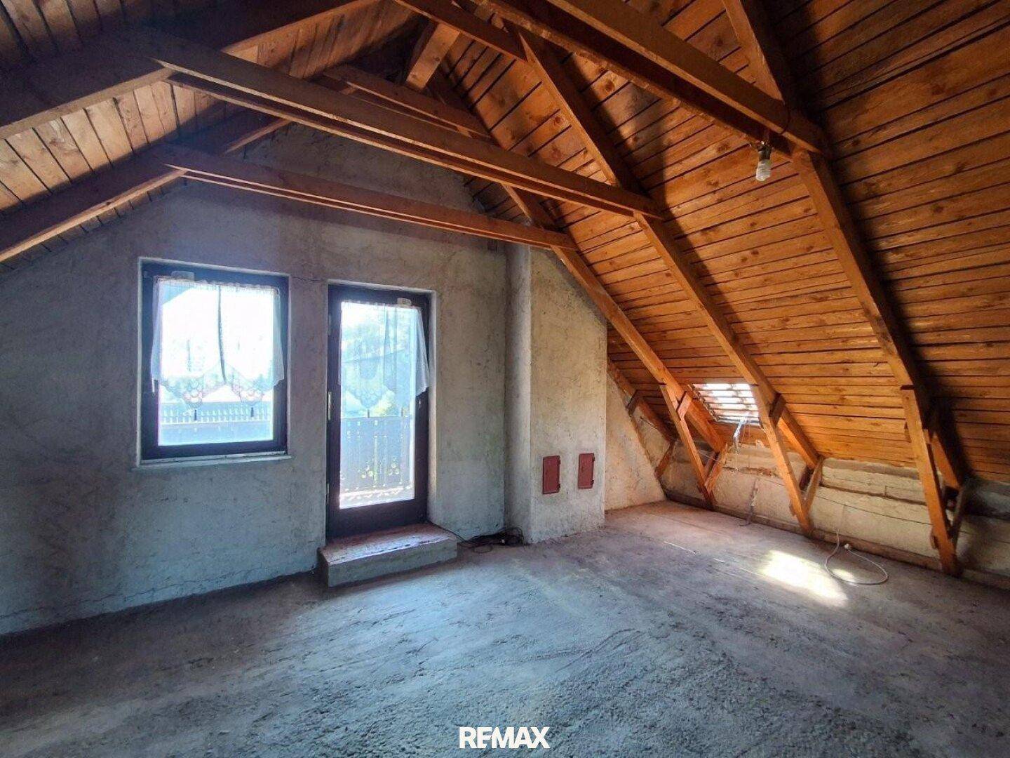 Unausgebauter Dachboden mit Holzbalken, Fenstern und Zugang zu einem möglichen Balkon oder Dachterrasse.