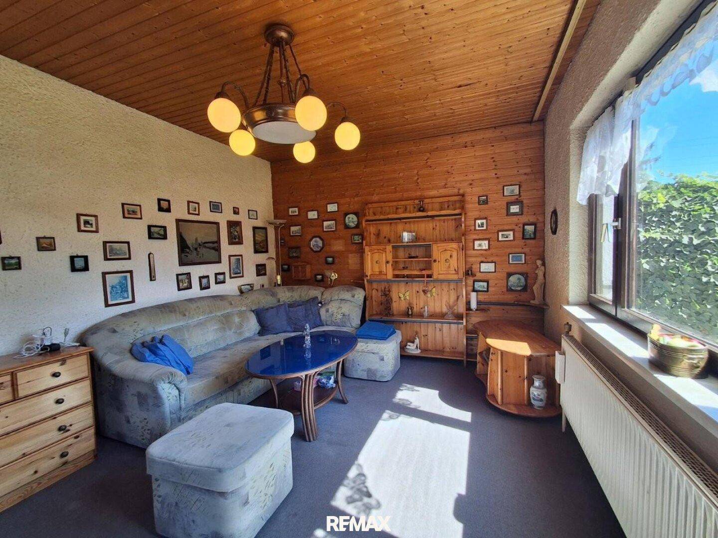 Geräumiges Wohnzimmer mit Sofa, Holzdecke und Fensterfront, die viel Licht und Blick ins Grüne bietet.
