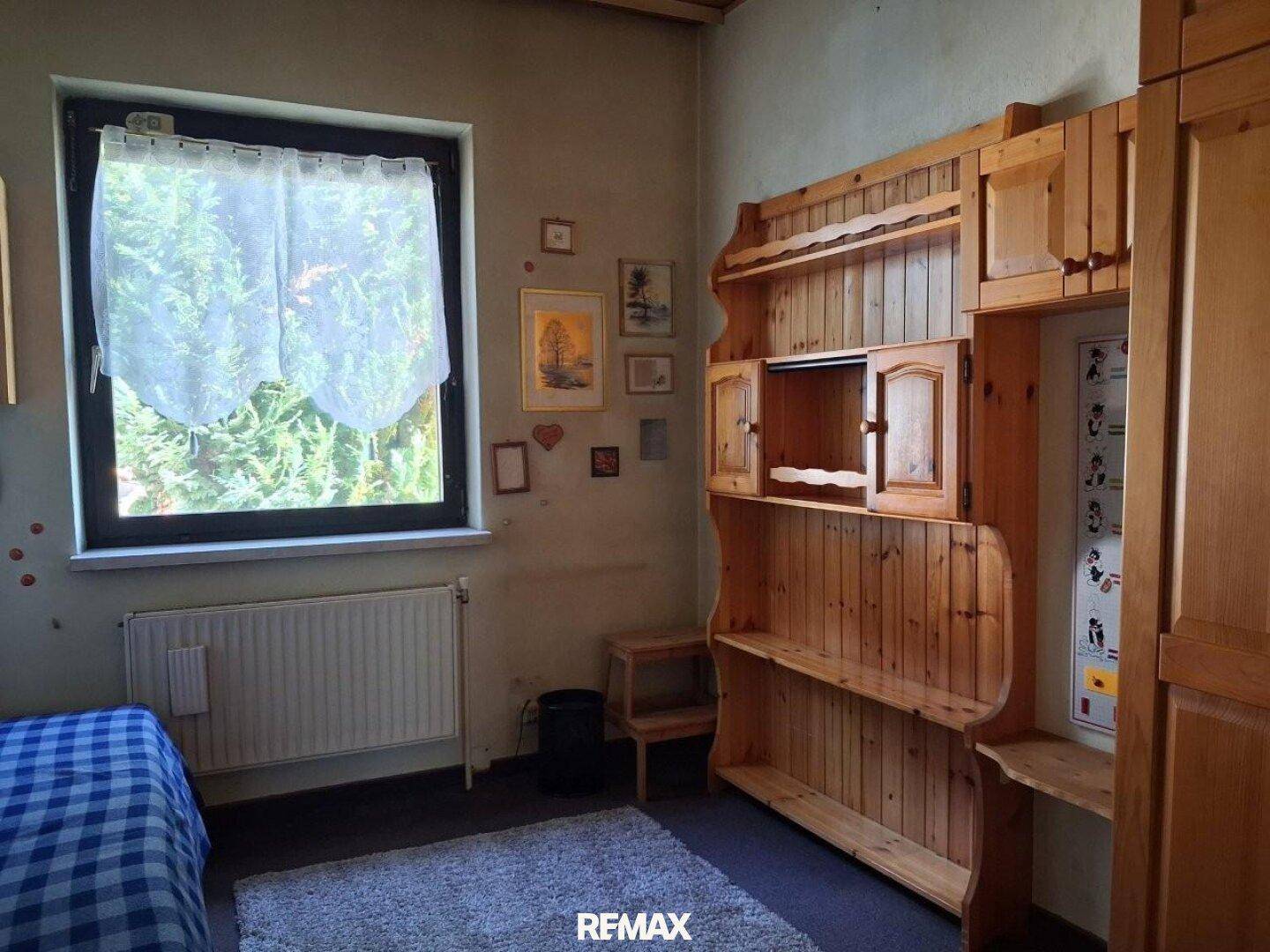 Helles Schlafzimmer mit Einzelbett, Holzregal und Fenster mit Blick ins Grüne, einfach eingerichtet.