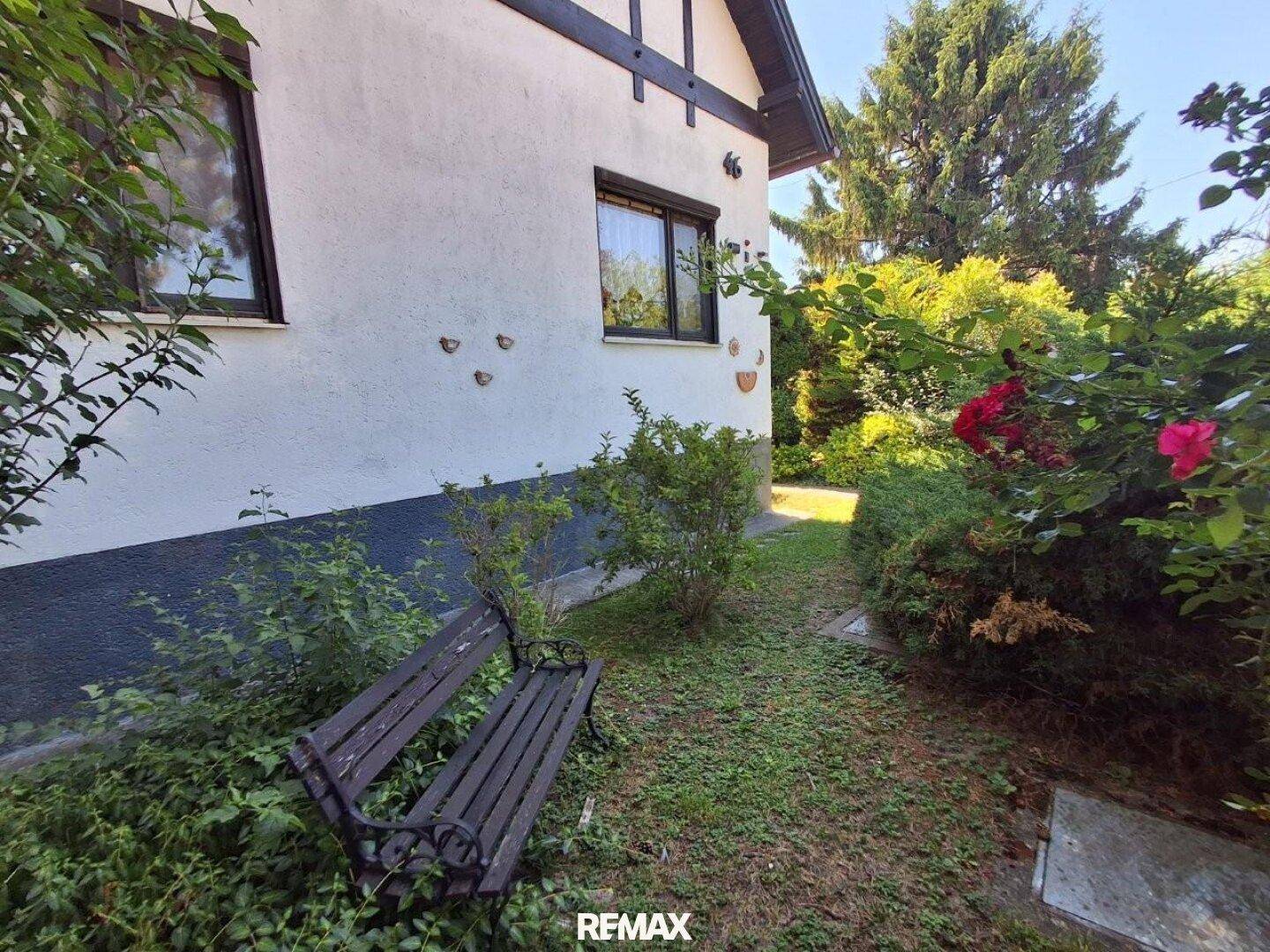 Gartenbereich neben dem Haus mit einer Holzbank und blühenden Rosen unter blauem Himmel.