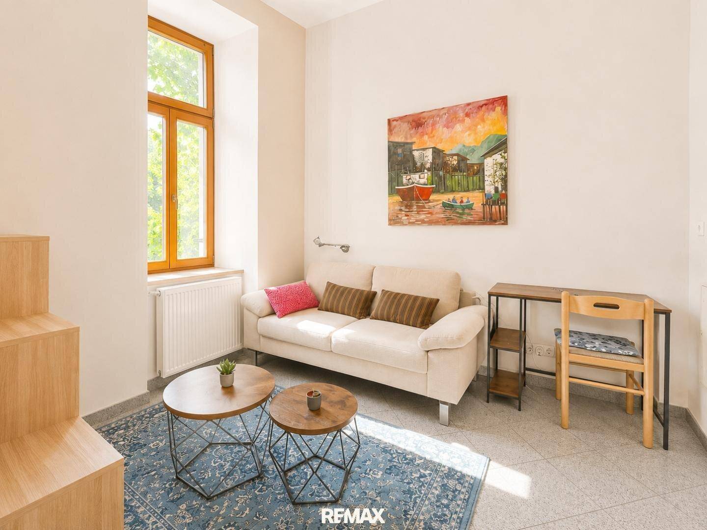 Gemütliches Wohnzimmer mit beigem Sofa, Teppich und einem kleinen Arbeitsbereich am Fenster.