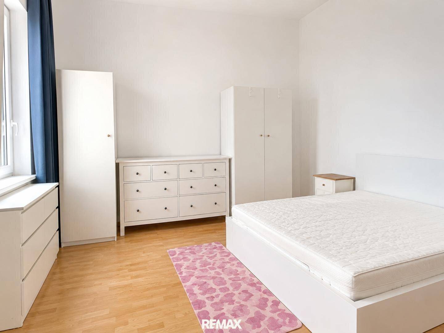 Schlafzimmer mit hellem Holzboden, weißen Möbeln und einem Fenster mit dunklen Vorhängen.