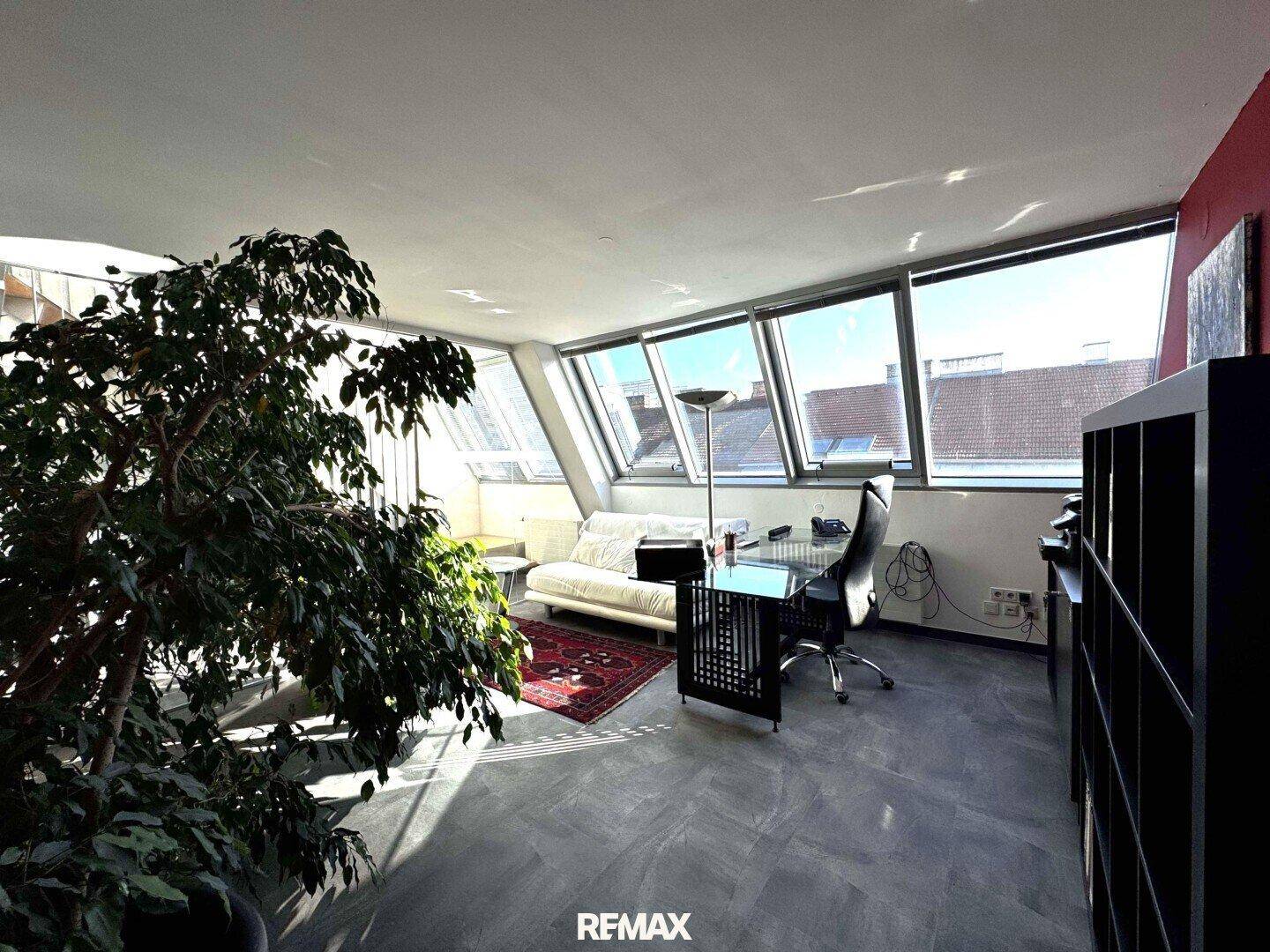 Geräumiges Büro mit viel Tageslicht durch die Dachfenster, einer großen Pflanze und Arbeitsbereich.
