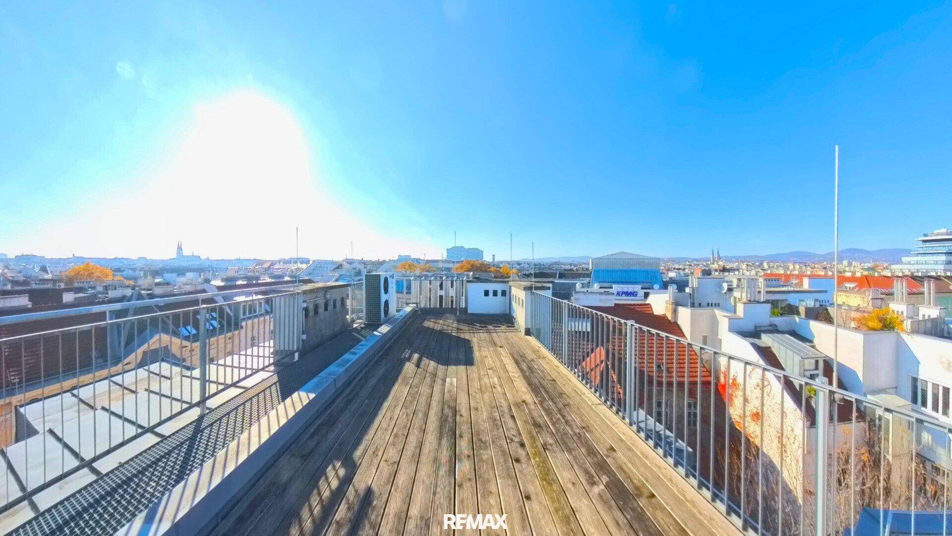 Geräumige Dachterrasse mit Holzbelag und weitem Blick über die Stadt an einem sonnigen Tag.