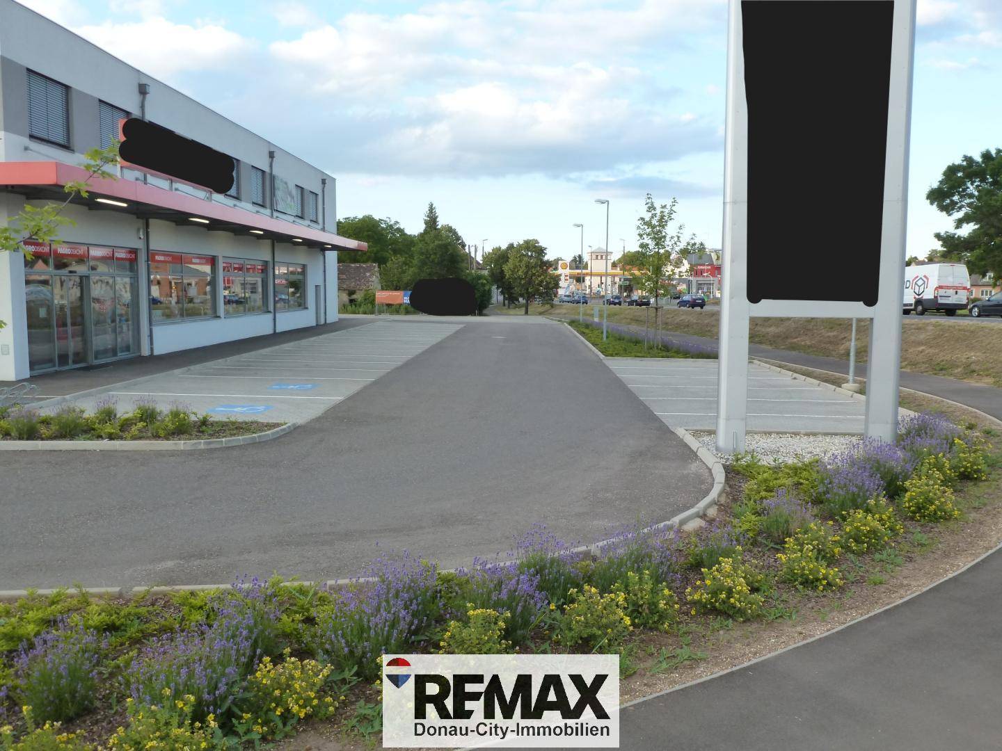 Geräumiger Parkplatz vor dem Geschäftsgebäude mit gepflegten Blumenbeeten und einer großen Werbetafel.