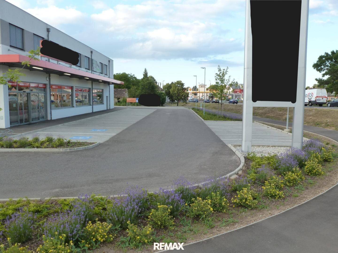 Ansicht des Parkplatzes vor dem Geschäftsgebäude mit gepflegten Blumenbeeten und einem großen Werbeschild.