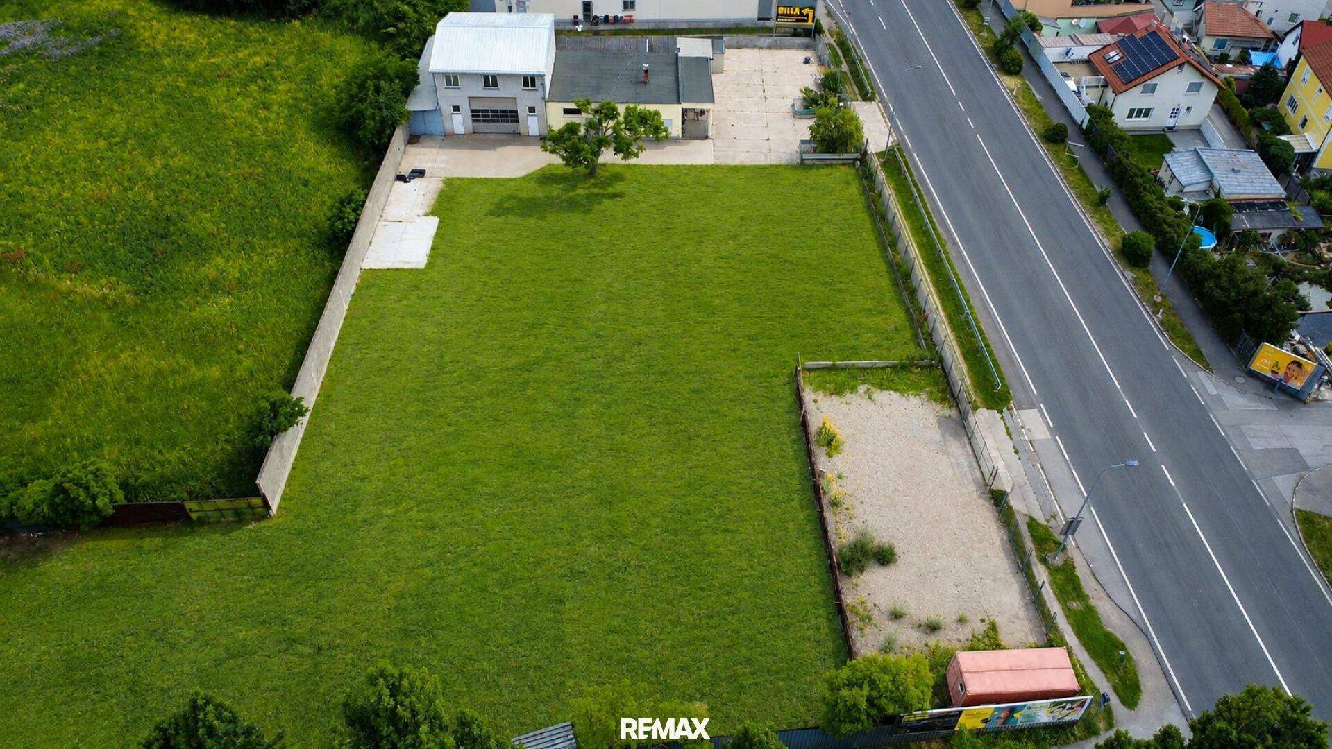 Überblick aus der Vogelperspektive über ein großes, grünes Grundstück neben einer Hauptstraße mit angrenzenden Gebäuden.