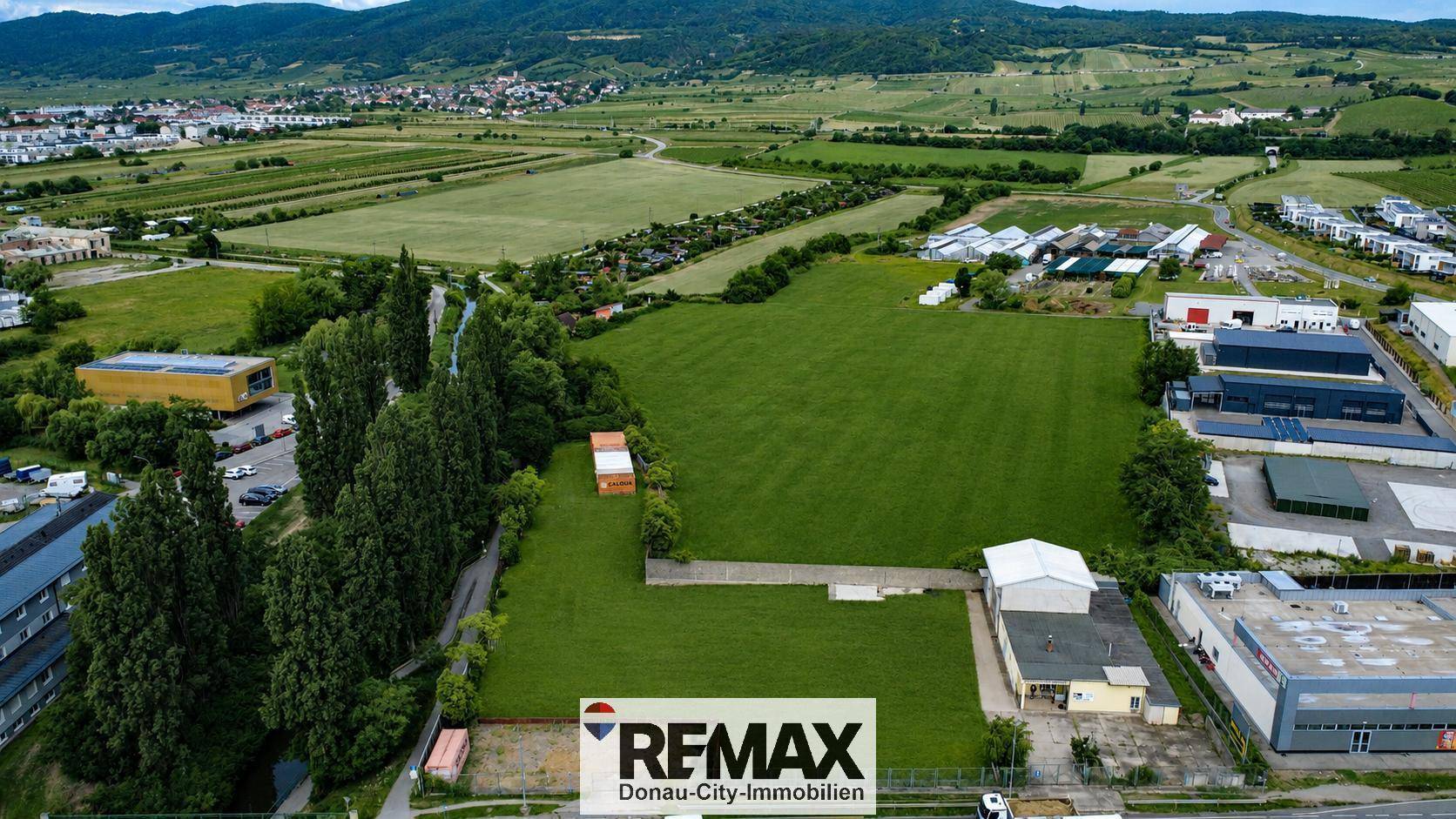 Panoramablick aus der Luft auf ein weitläufiges grünes Grundstück mit Bergen im Hintergrund und umliegender Bebauung.