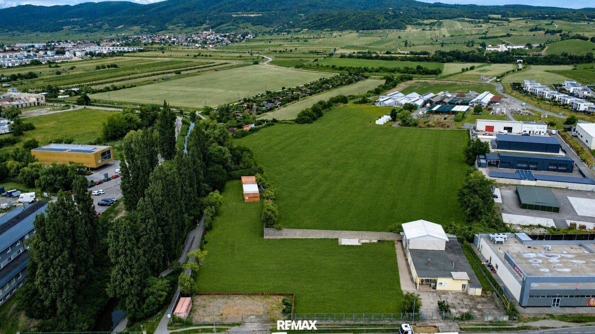 Panoramische Luftaufnahme eines weitläufigen Grüngeländes mit Bergen im Hintergrund und städtischer Bebauung.