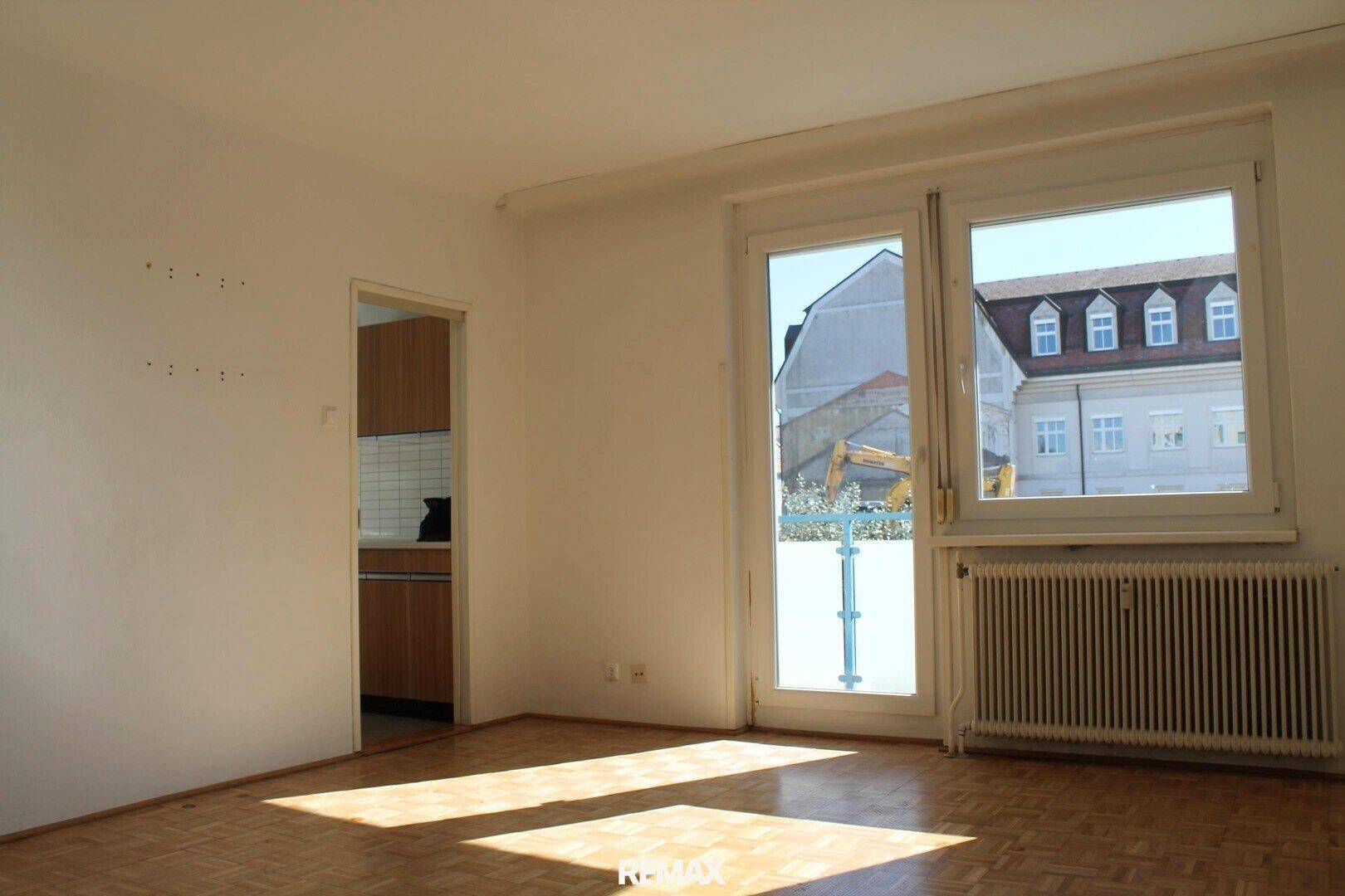 Lichtdurchflutetes Wohnzimmer mit Parkettboden, Heizkörper und Blick auf die umliegenden Gebäude.