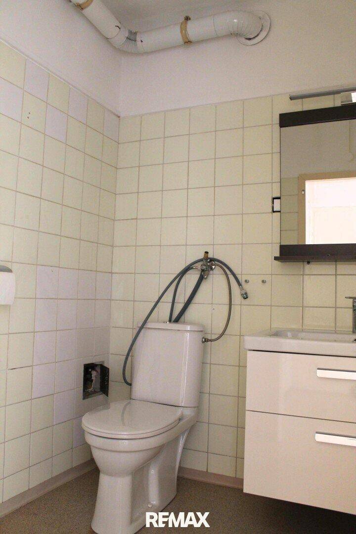 Badezimmer mit Toilette, Einzelwaschbecken und hellbeigen Fliesen, mit sichtbaren Rohrleitungen.