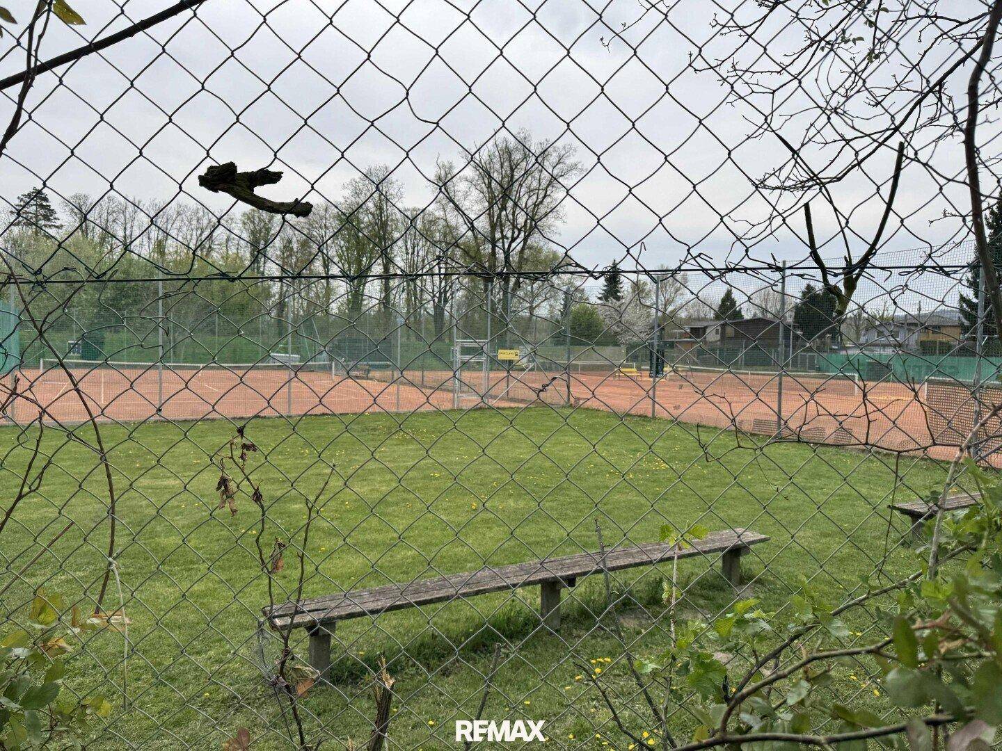 Blick durch einen Zaun auf zwei rote Tennisplätze mit einer Holzbank im Vordergrund.